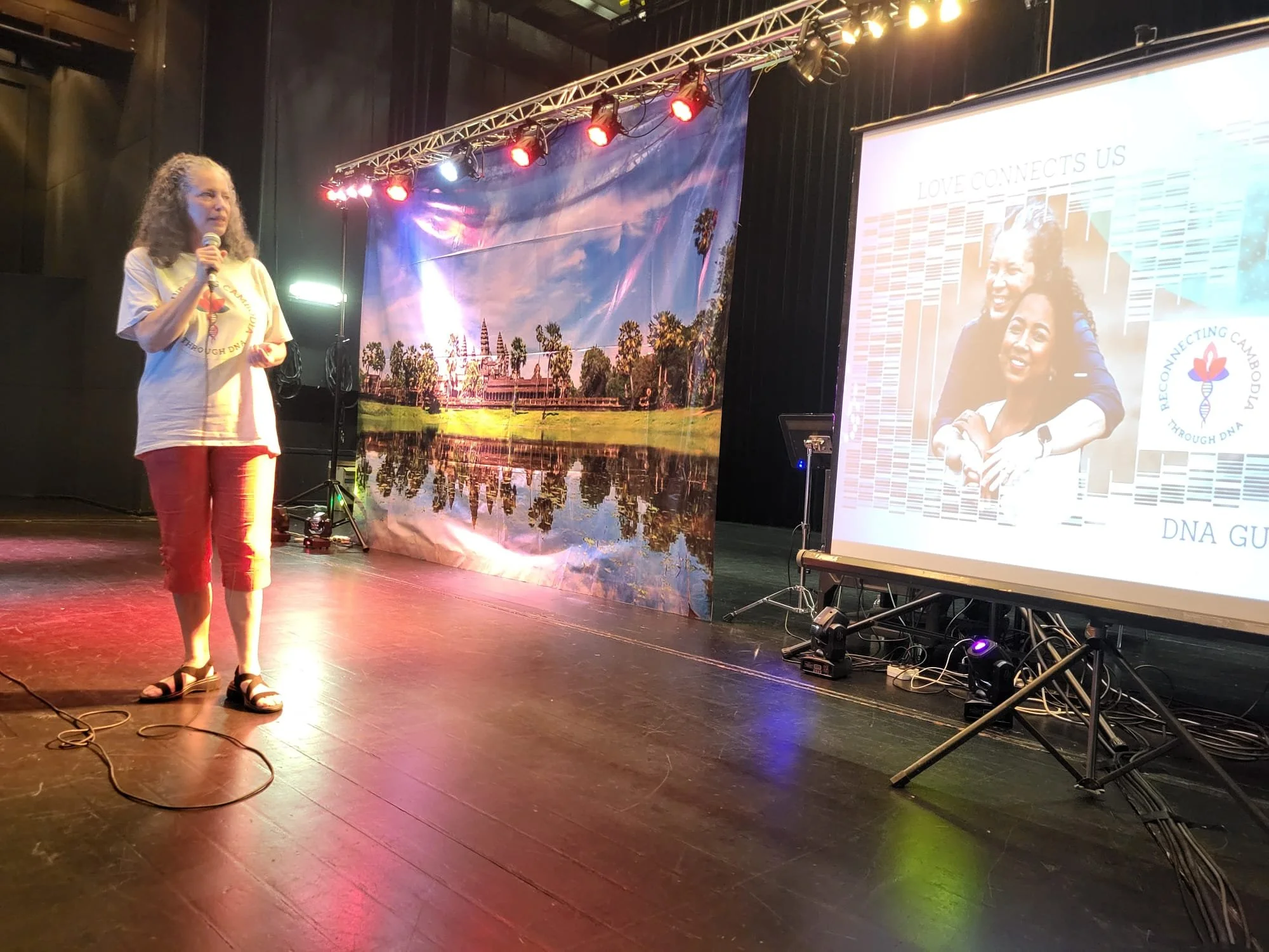 A woman with grey hair stands on a stage speaking into a microphone. Behind her is a large backdrop depicting the Angkor Wat temple, and to her right is a projector screen displaying a slide titled "LOVE CONNECTS US" with a photo of two women embeace