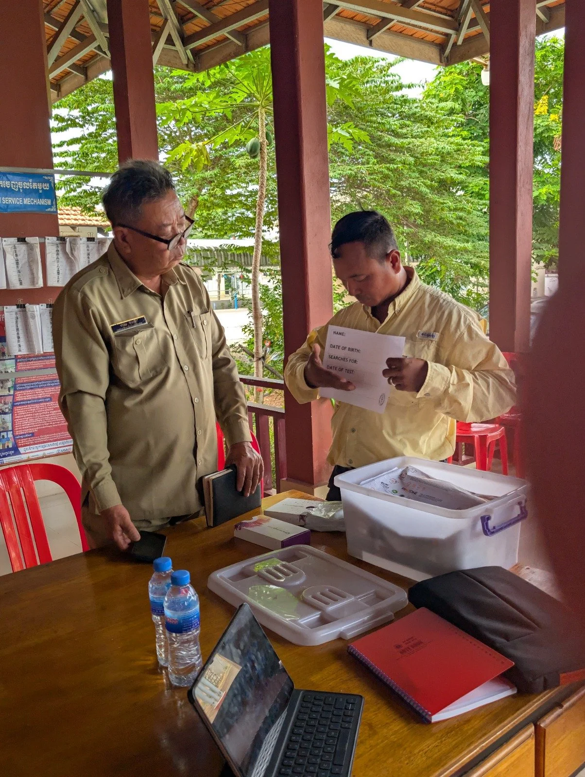 Two men in beige uniforms standing at a table, examining documents, in a setting with a wooden roof and lush green trees outside. There are water bottles, laptops, and office supplies on the table.