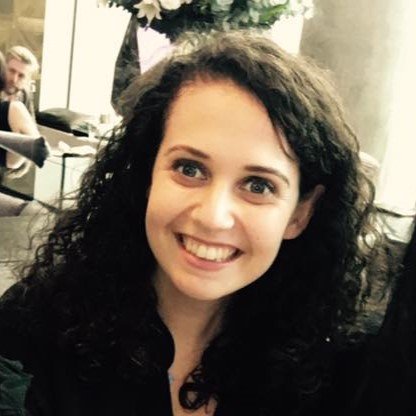 A young woman with long curly hair smiling in an indoor setting, with other people and floral arrangements in the background.