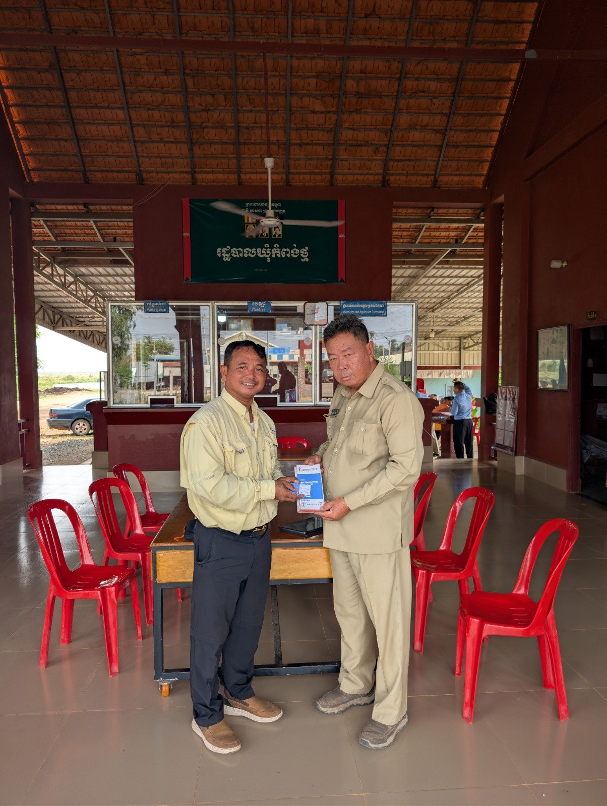 Two men in khaki uniforms exchanging a document or an item inside a building with red chairs and a counter in the background.