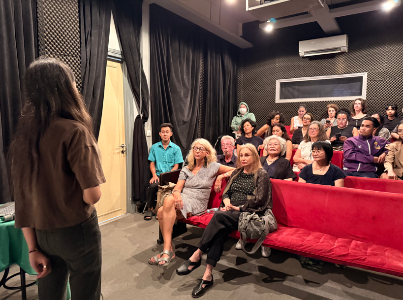 A woman is speaking in front of a group of people in a small theater or conference room with black soundproof walls and red seats. The audience, diverse in age and gender, is seated and attentively listening.