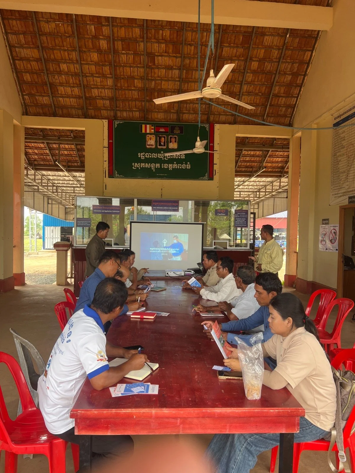 A group of people sitting at a long conference table in a pavilion, attending a presentation with a projector and screen. Several individuals are reading pamphlets or taking notes, and a man is standing near the screen.