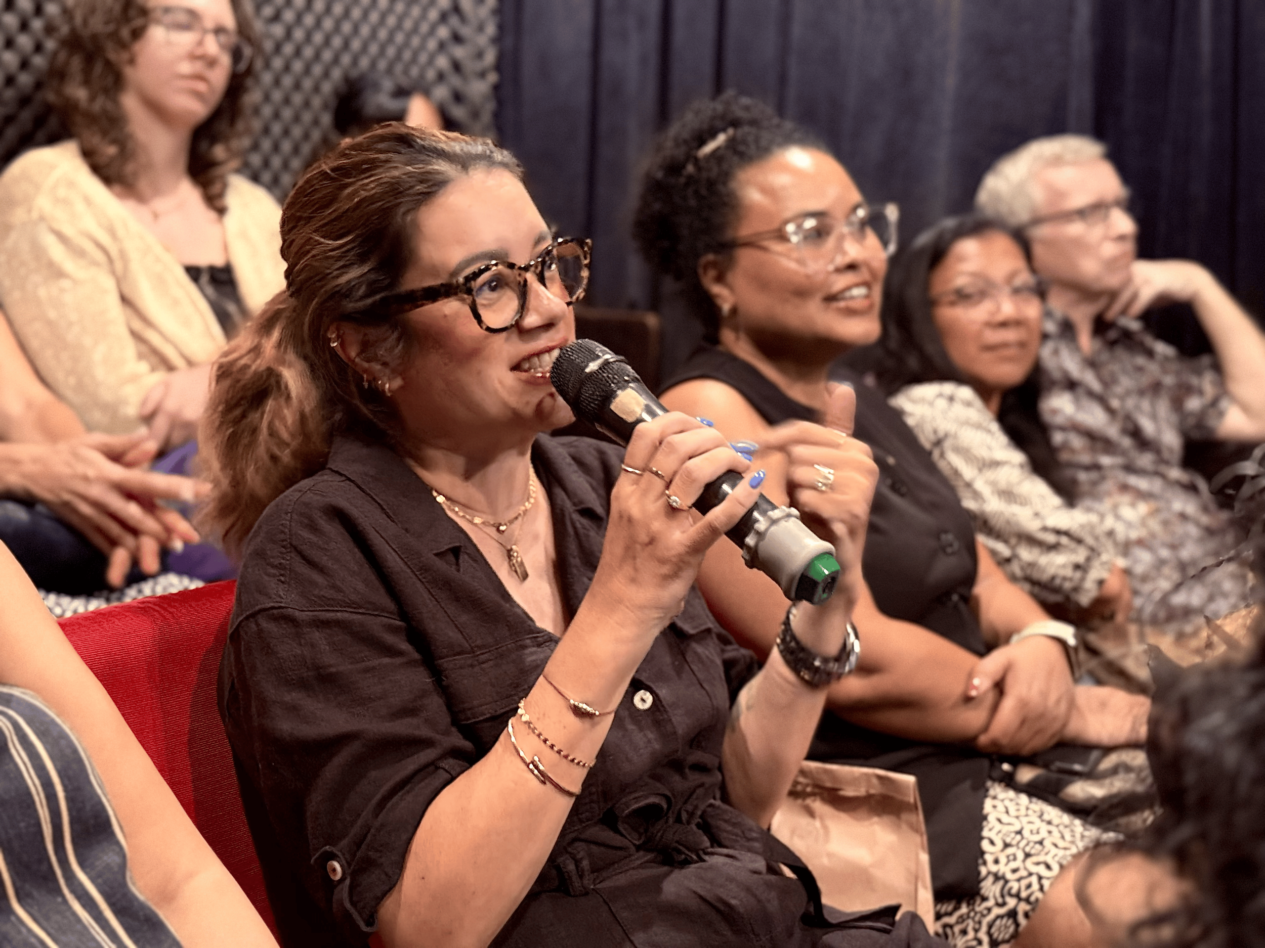 A woman with glasses holding a microphone speaking at an event, seated among other women in an auditorium.