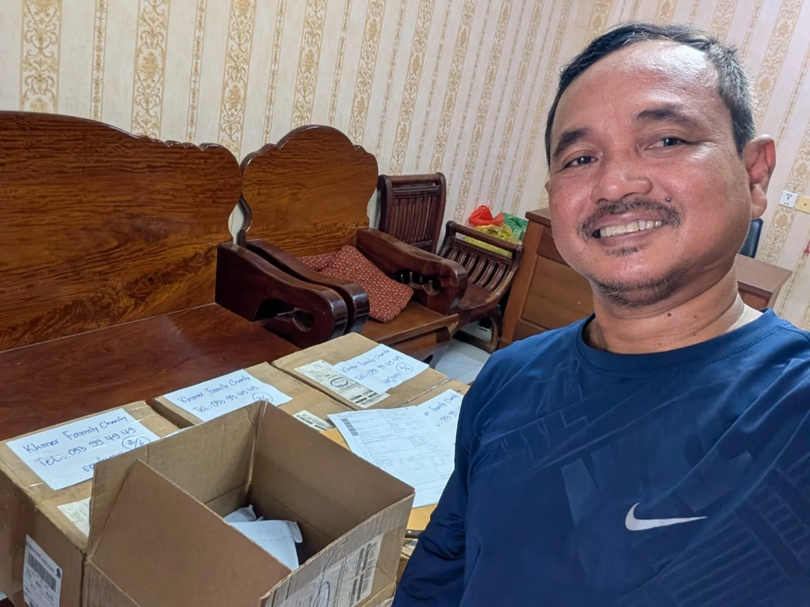A man smiling and taking a selfie in a room with wooden furniture and packages labeled 'Khmer Family Church' on a table.