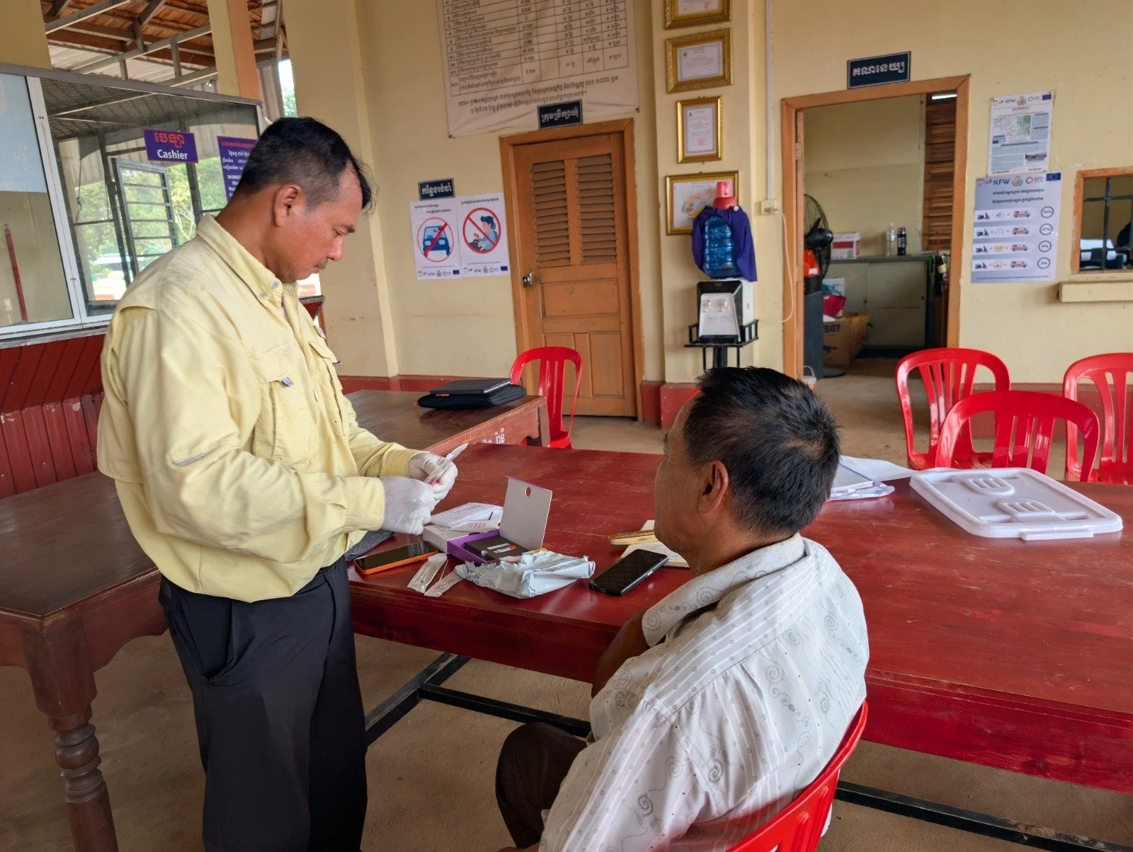 A man in a yellow shirt and a man in a white striped shirt sitting at a wooden table with a red plastic chair. A person wearing gloves standing, possibly filling out paperwork or examining documents. The setting appears to be a small office or recept