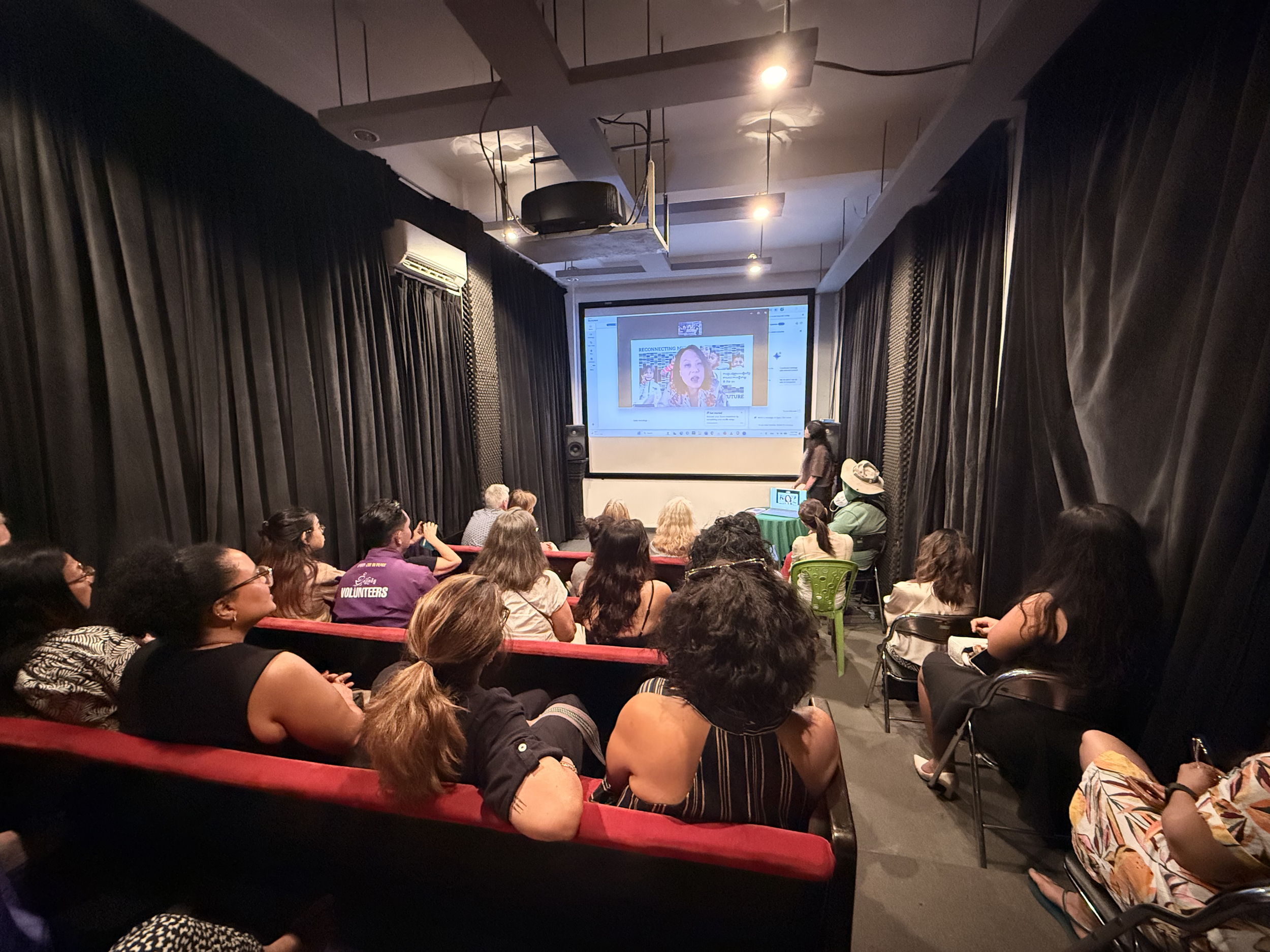 A large audience seated in a theater or conference room, watching a presentation on a large screen at the front. The room has black curtains on both sides and a woman standing in front of the audience near a laptop.