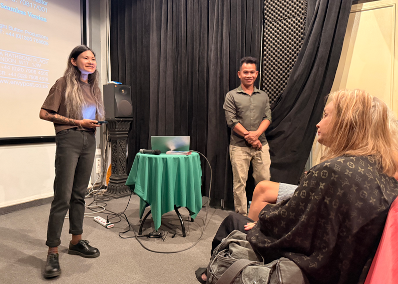A presentation or speech taking place in a room with black curtains, a projector screen, and a small green table with a laptop. A woman with long hair and dark clothing is speaking, while a man in a grey shirt stands nearby. An audience member with b
