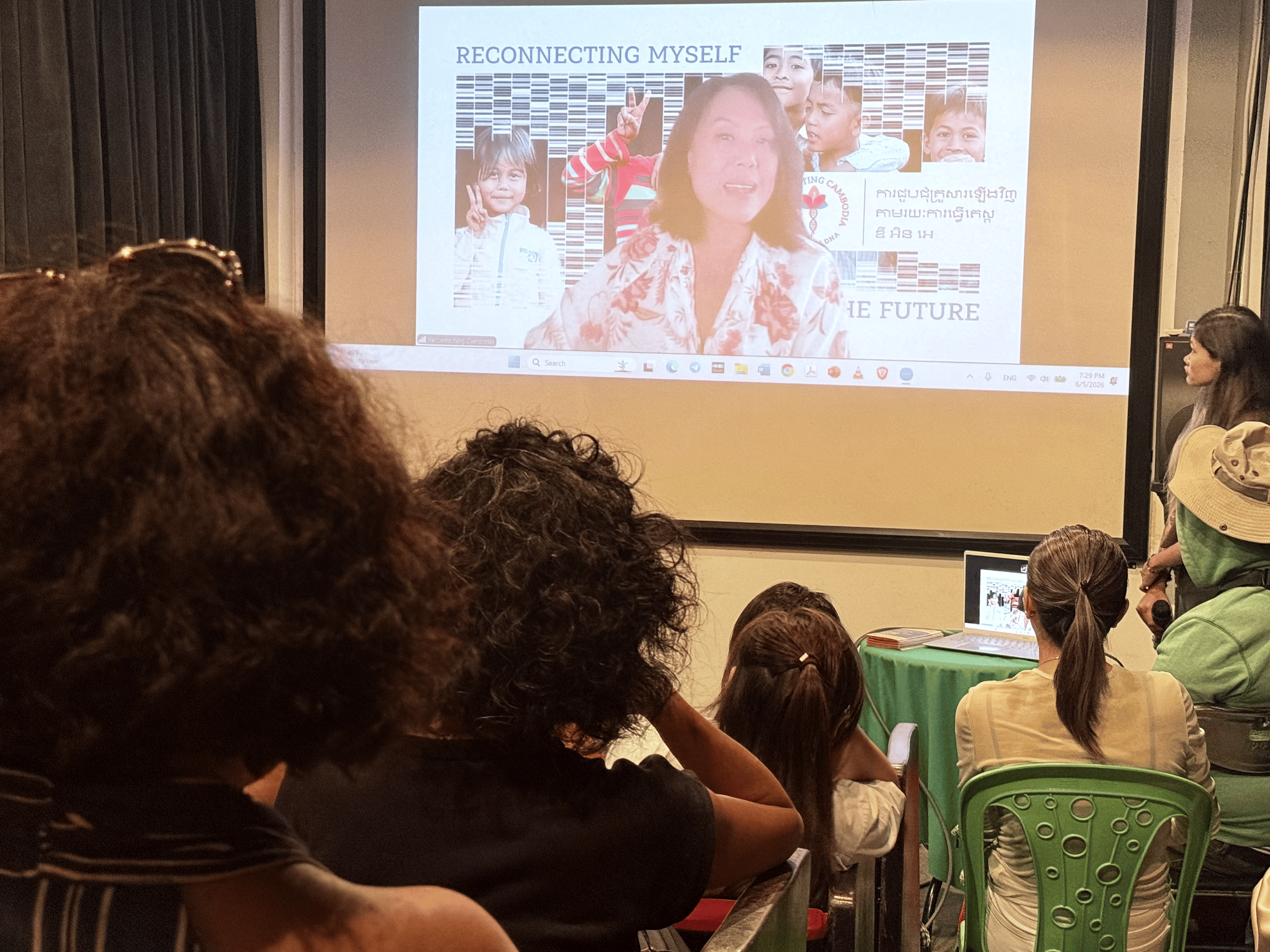 A presentation slide titled 'Reconnecting Myself' featuring a woman and several children, with a diverse audience attending in a conference room, some seated and one standing with a laptop.