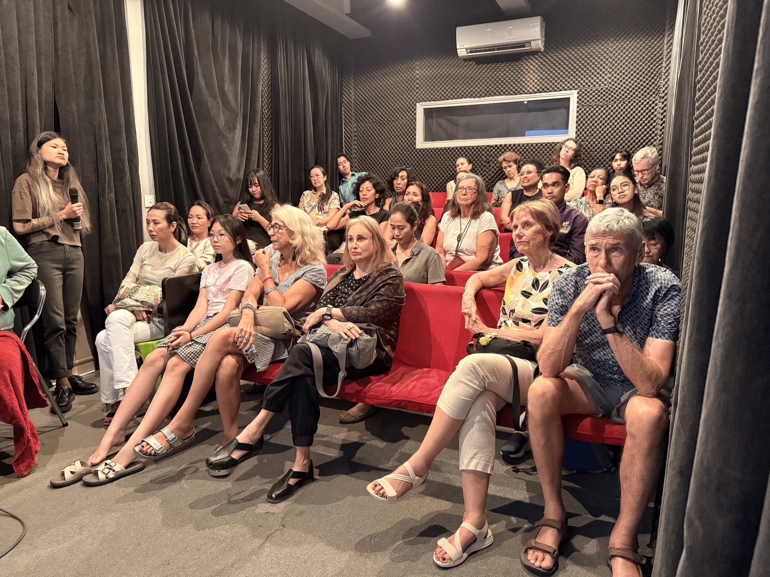 An audience seated in red theater seats, attentively listening to a woman standing with a microphone in a dark, soundproofed room with black acoustic foam panels, curtains, and a small window.