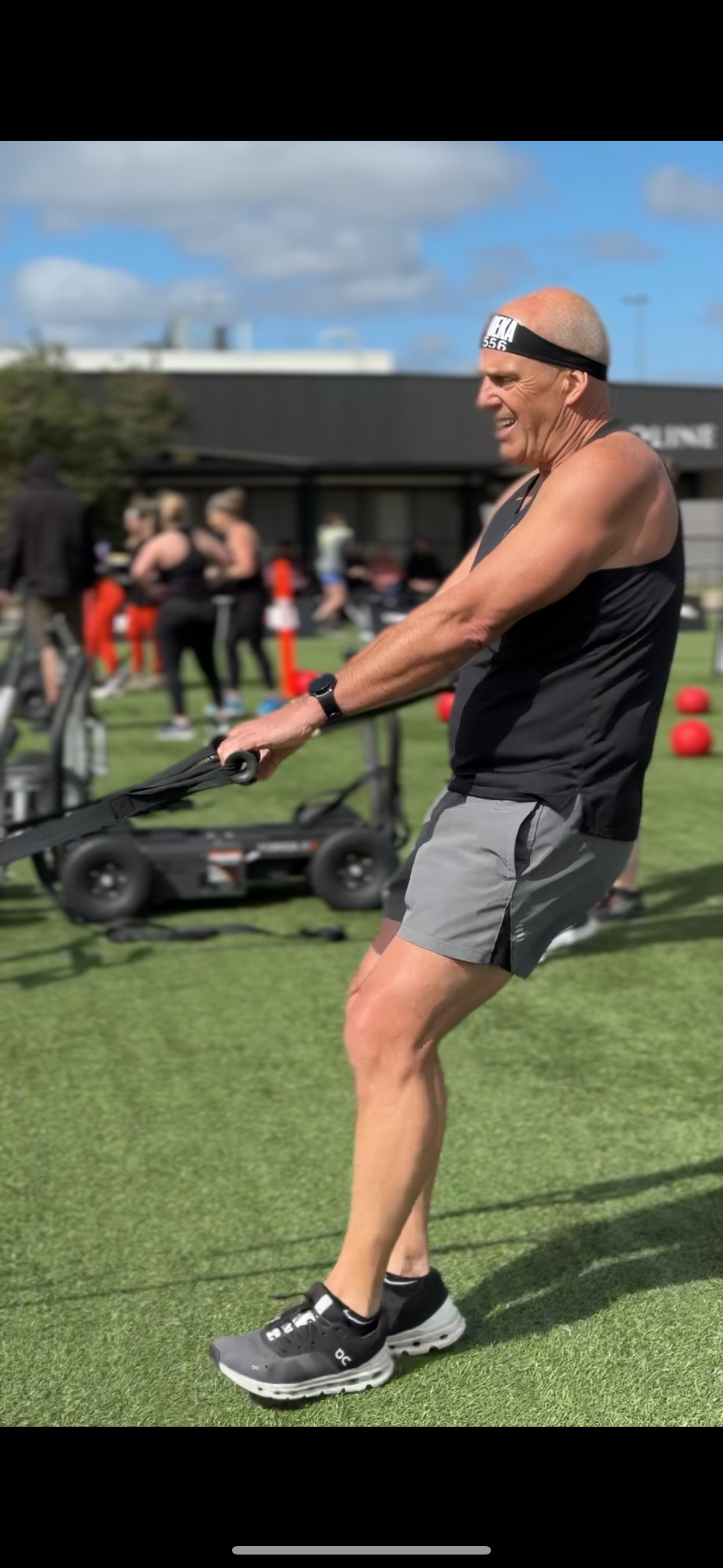 Man exercising outdoors with resistance bands, wearing a black tank top, gray shorts, and a headband.