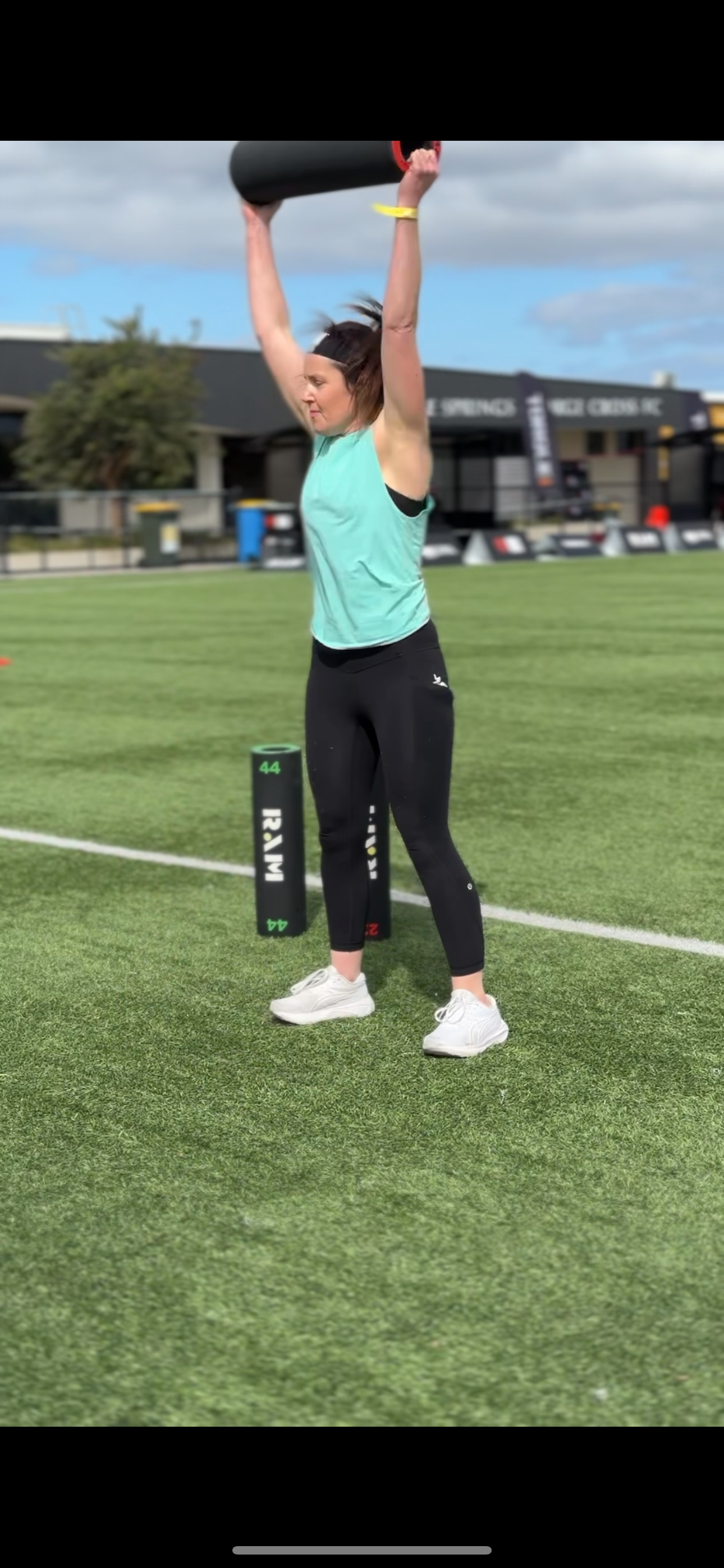 Woman lifting a black exercise log overhead on a sports field.