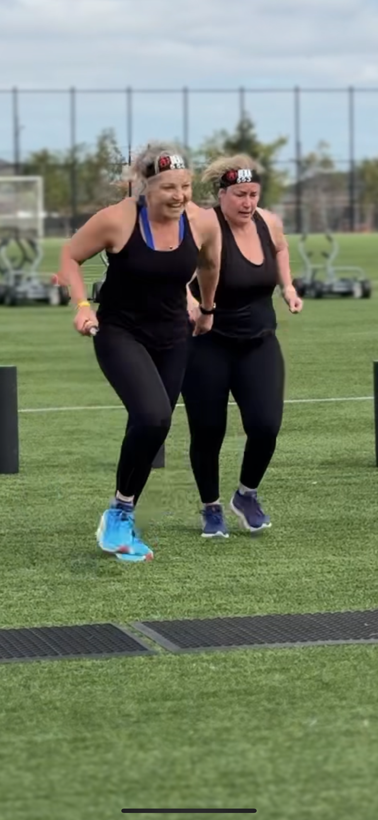 Two women wearing black workout clothes and headbands race each other on a grassy field, with exercise equipment in the background.