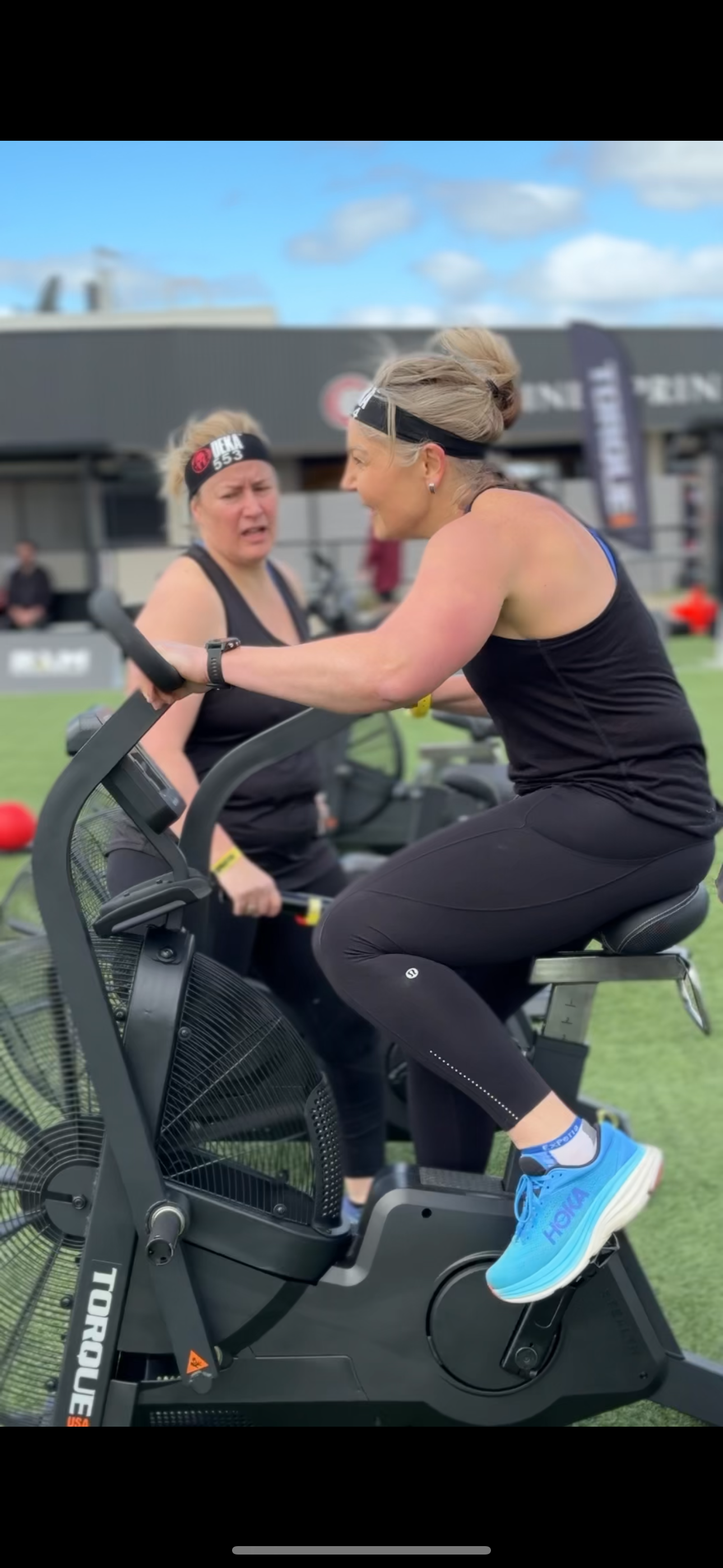 Two women in athletic wear engage in conversation. One is seated on an air bike, and they appear to be participating in an outdoor fitness event.