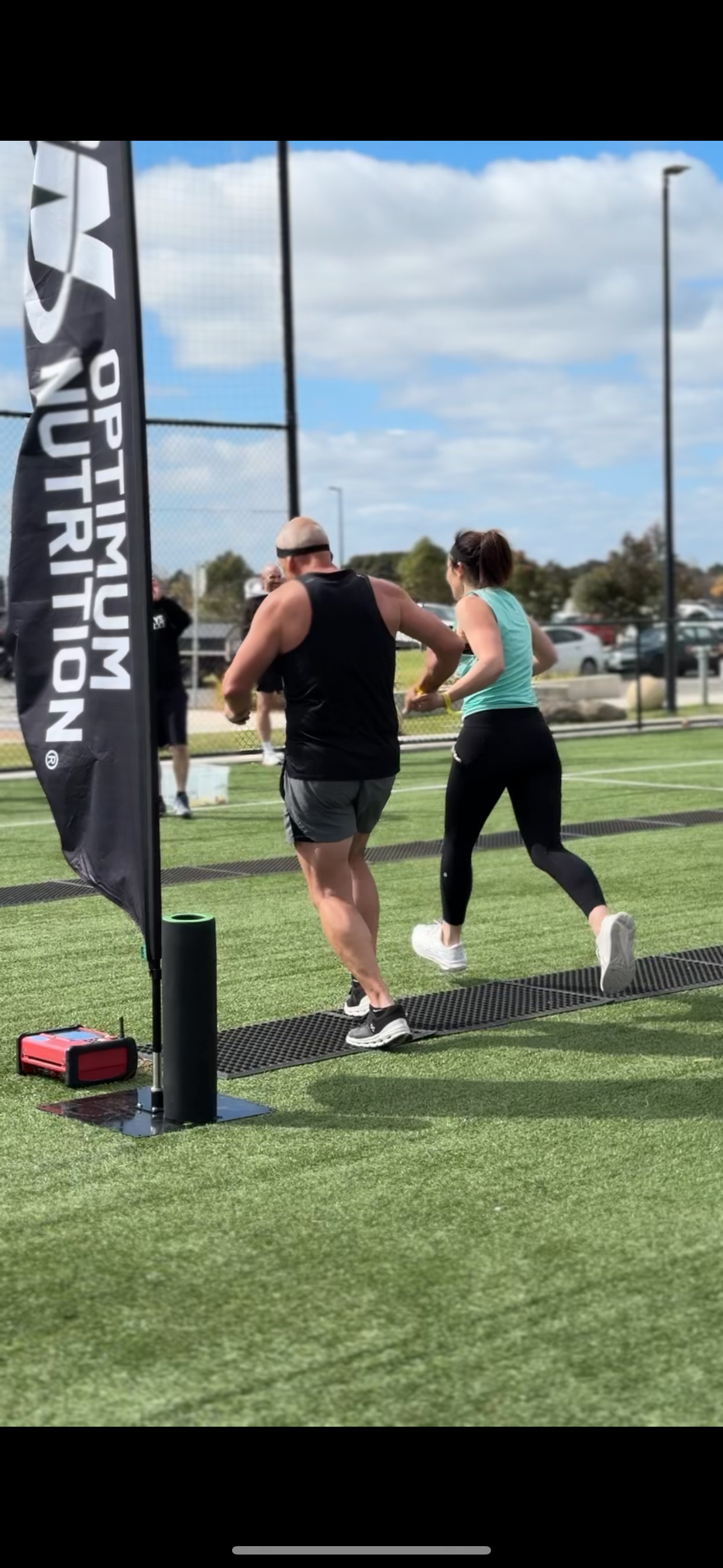 Two people running on a grassy field beside an Optimum Nutrition banner.