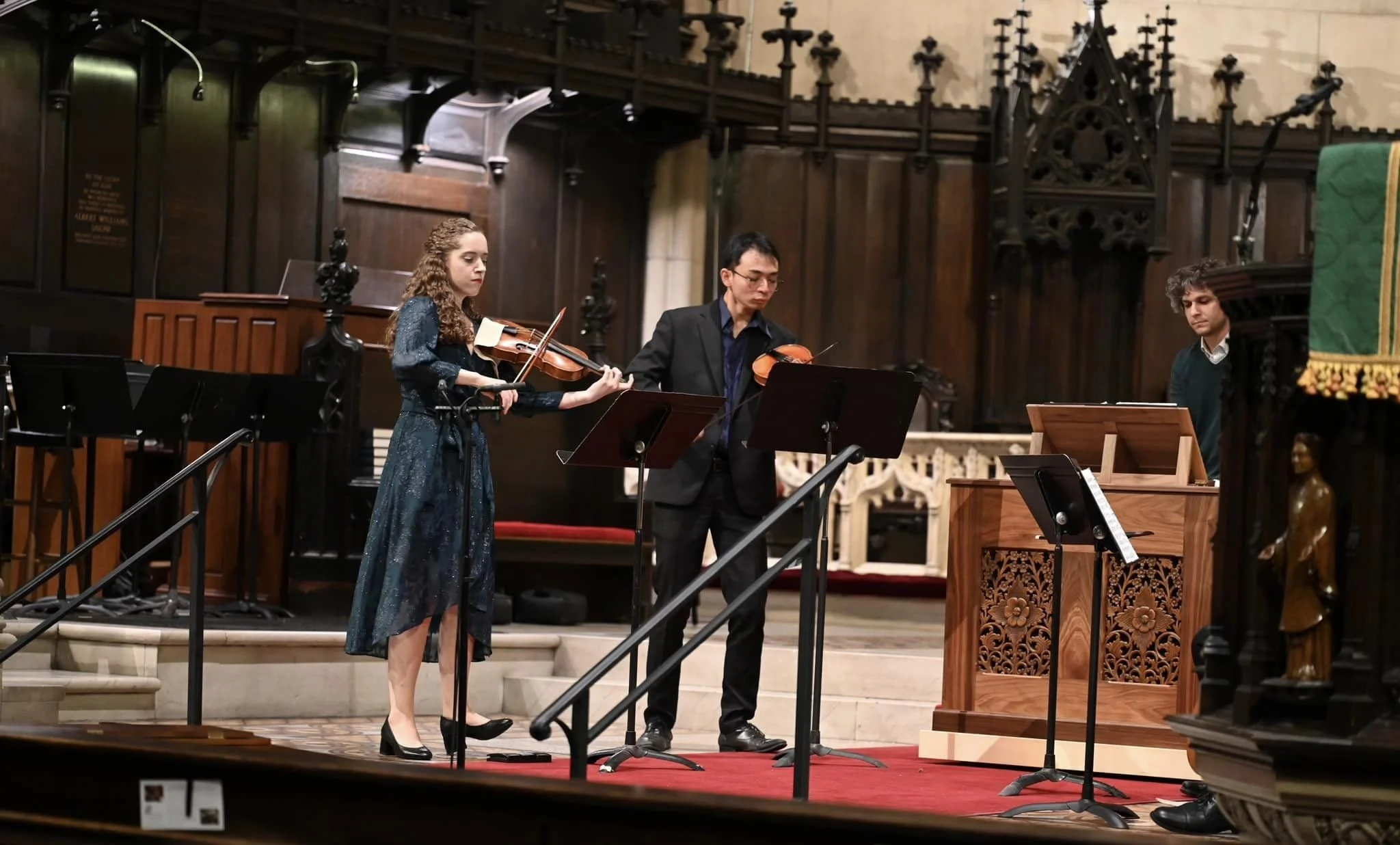 Three musicians performing in a church or cathedral: a woman playing the violin, a man playing the viola, and another man playing the piano. The setting features ornate wooden architecture and gothic-style decor.