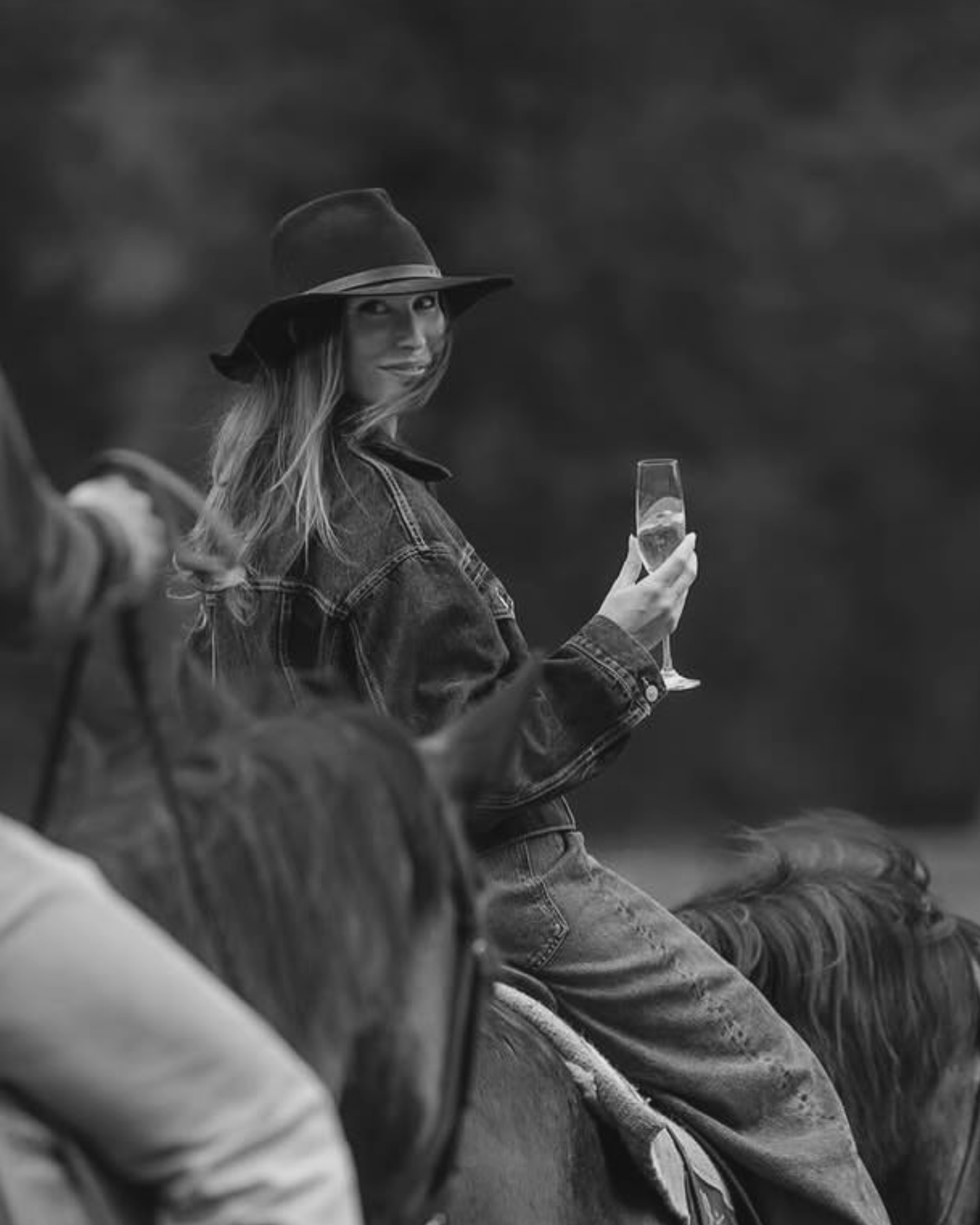 A woman wearing a wide-brimmed hat and denim jacket, holding a glass of champagne, sitting outdoors on grass.