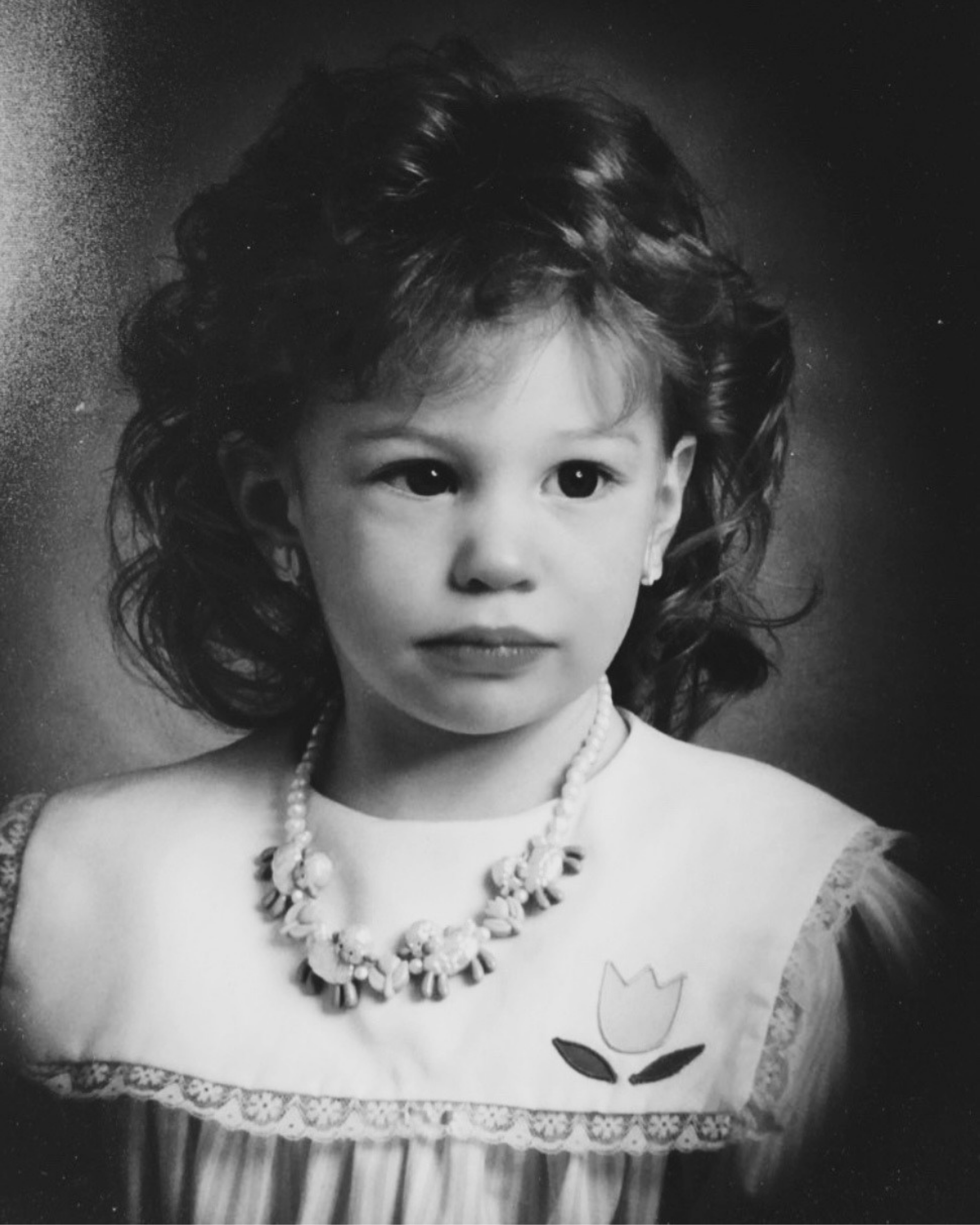 Black-and-white portrait of a young girl with dark, wavy hair, wearing pearl jewelry and a lace dress, holding a handmade card with a flower drawing.