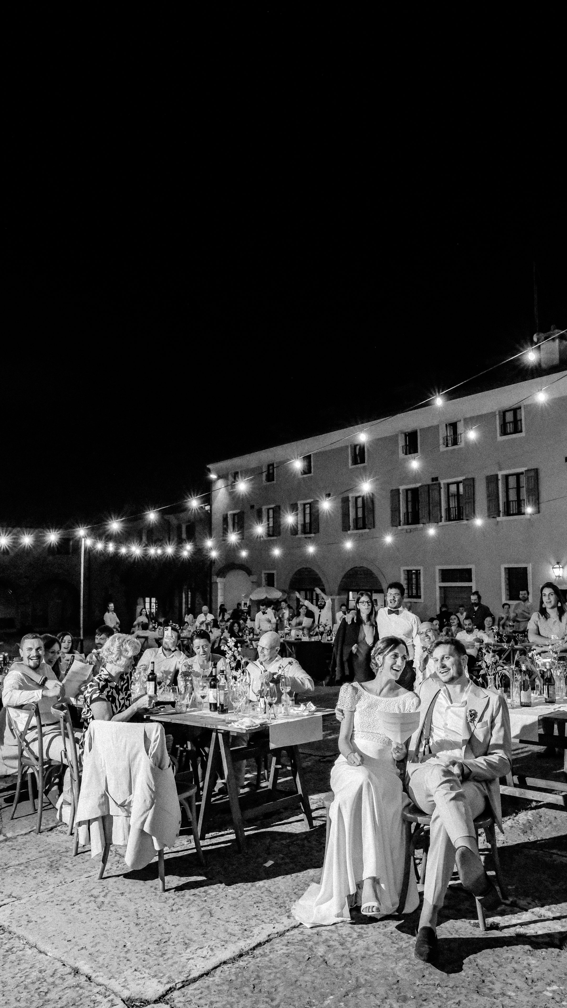 Nela and Davide sitting together among their family and friends during their outdoor wedding dinner beneath string lights in the courtyard of Serego Alighieri near Verona.