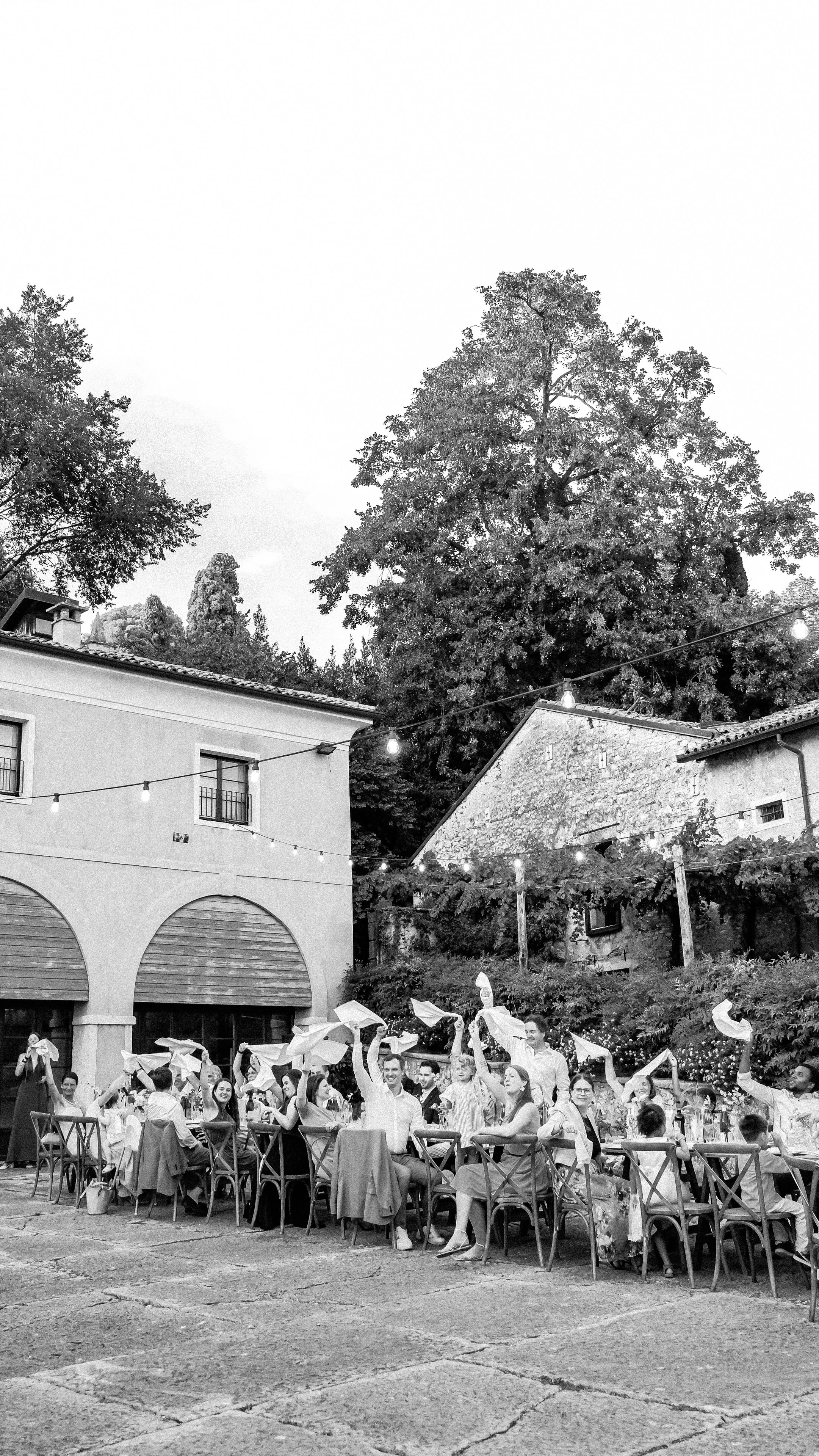 Wedding guests waving white napkins in the air during an outdoor dinner under string lights in the historic courtyard of Serego Alighieri.