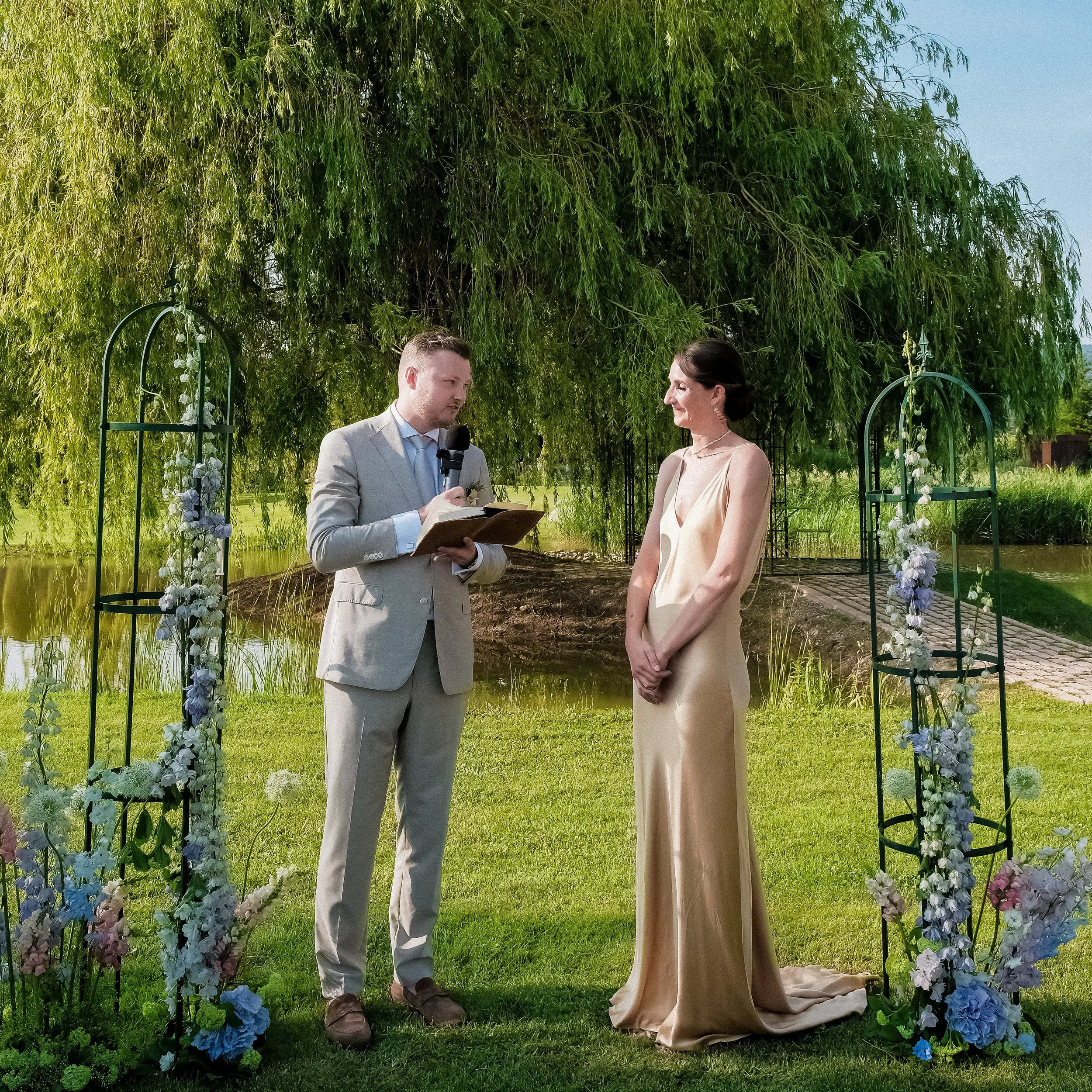 Outdoor wedding ceremony at Domaine des Andéols in Provence, groom reading vows to bride in a gold dress by the lake.