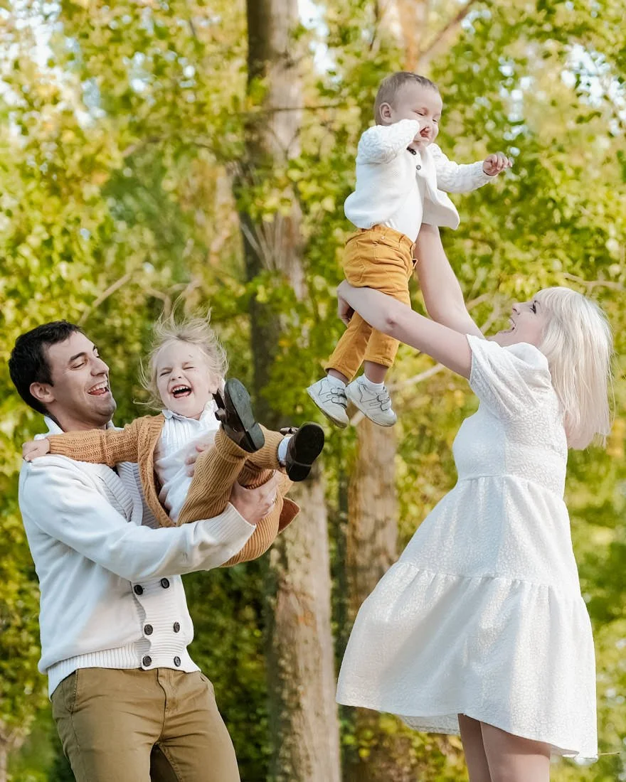 Joyful multicultural family moment — parents lifting their children during an outdoor session surrounded by natural light and trees.