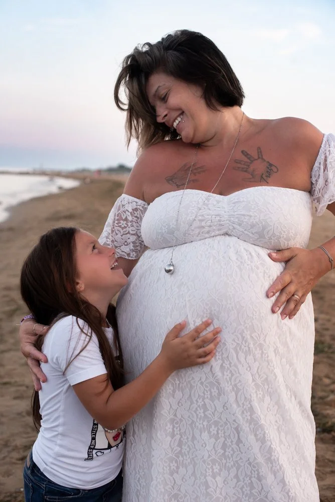 Una donna incinta con una maglietta bianca in pizzo e una collana con una palla, che sorride mentre abbraccia una bambina con camicia bianca. Sono sulla spiaggia al tramonto, si sorridono e si toccano la pancia.