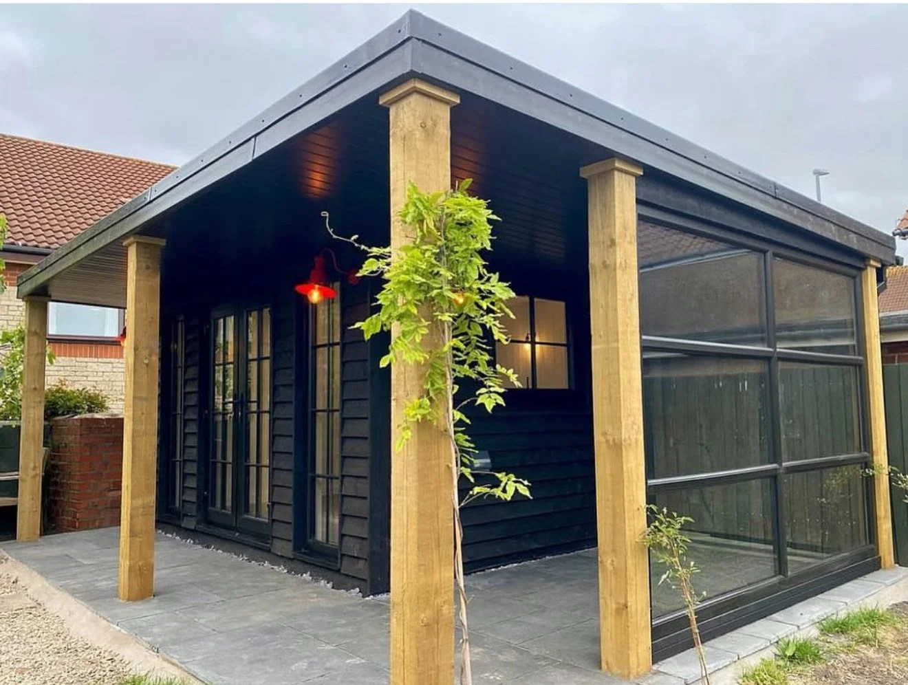 A modern outdoor garden room with black wooden walls, large glass windows, and a black metal roof supported by wooden posts, with a small tree and outdoor lighting.