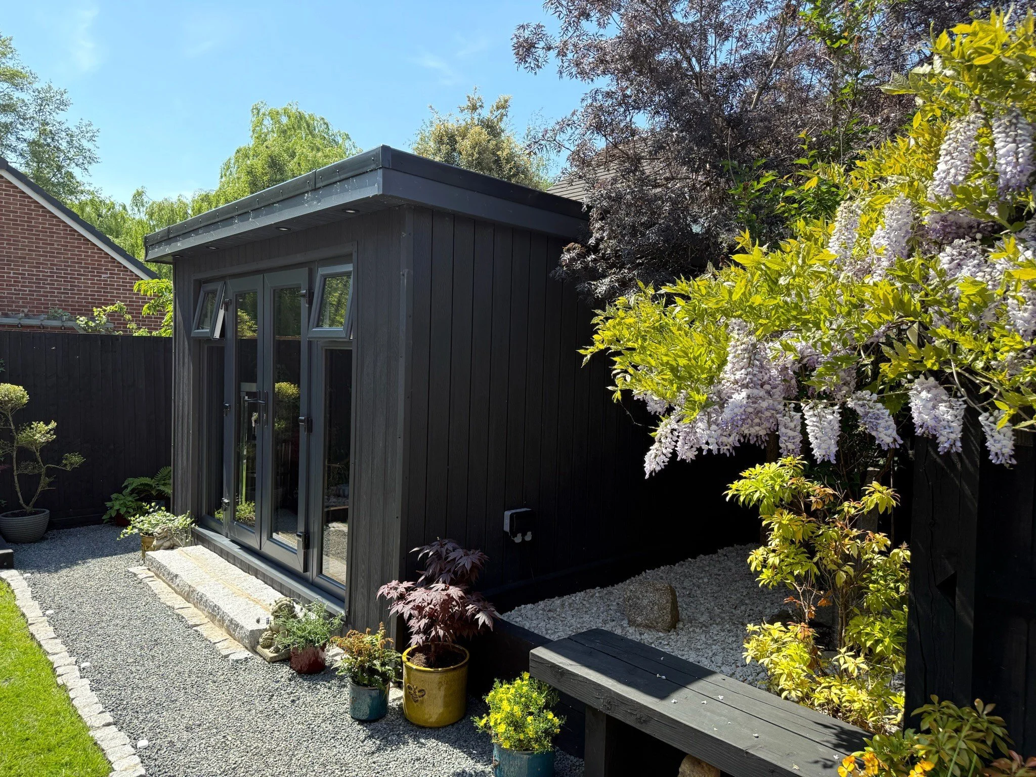 Black composite gardeb room with glass double doors and two slim windows on top situated in a landscaped garden with wisteria and potted plants, flowering bushes, and gravel pathway, under a clear blue sky.