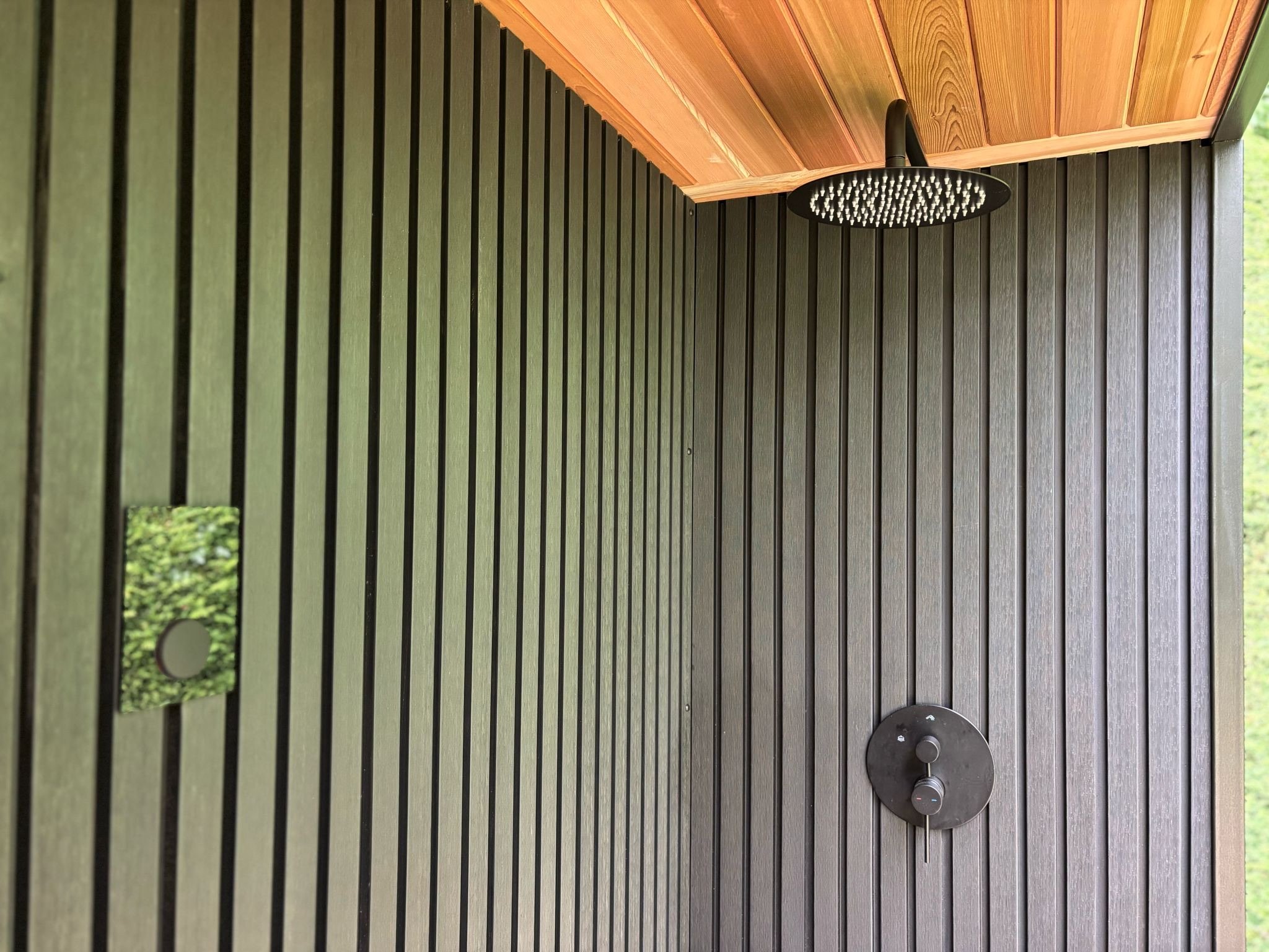 Outdoor shower with black showerhead and control knobs, surrounded by black composite vertical cladding and a cedar roof.