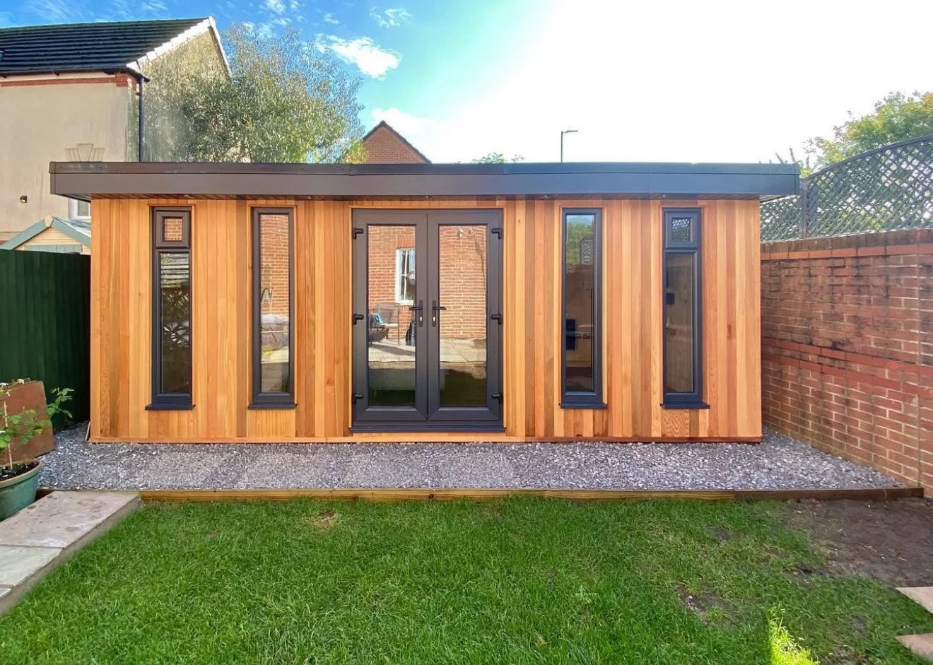 A cedar garden rooms with four full height feature windows and double doors, situated in a backyard with green grass, gravel, and a brick wall.