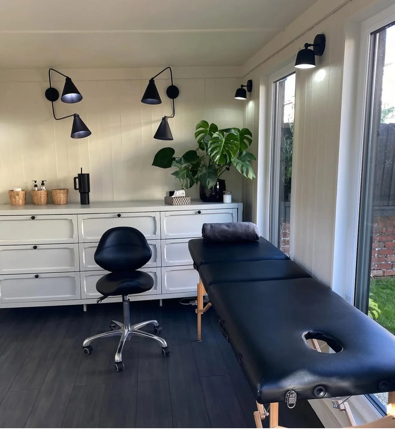 A garden studio / treatment room with a black massage table, a black office chair, white cabinet with baskets, a green leafy plant, and black wall-mounted lamps, with large windows showing an outdoor view.