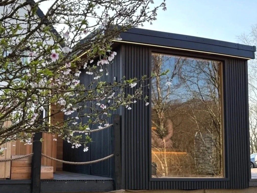 A mobile sauna with large picture window, black vertical panel siding, and a wooden deck surrounded by trees and blooming flowers.