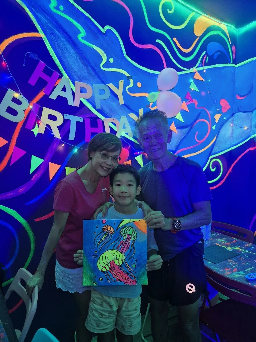 Family of three celebrating birthday in a glow-in-the-dark room with neon decorations and jellyfish artwork, holding a colorful drawing of jellyfish.