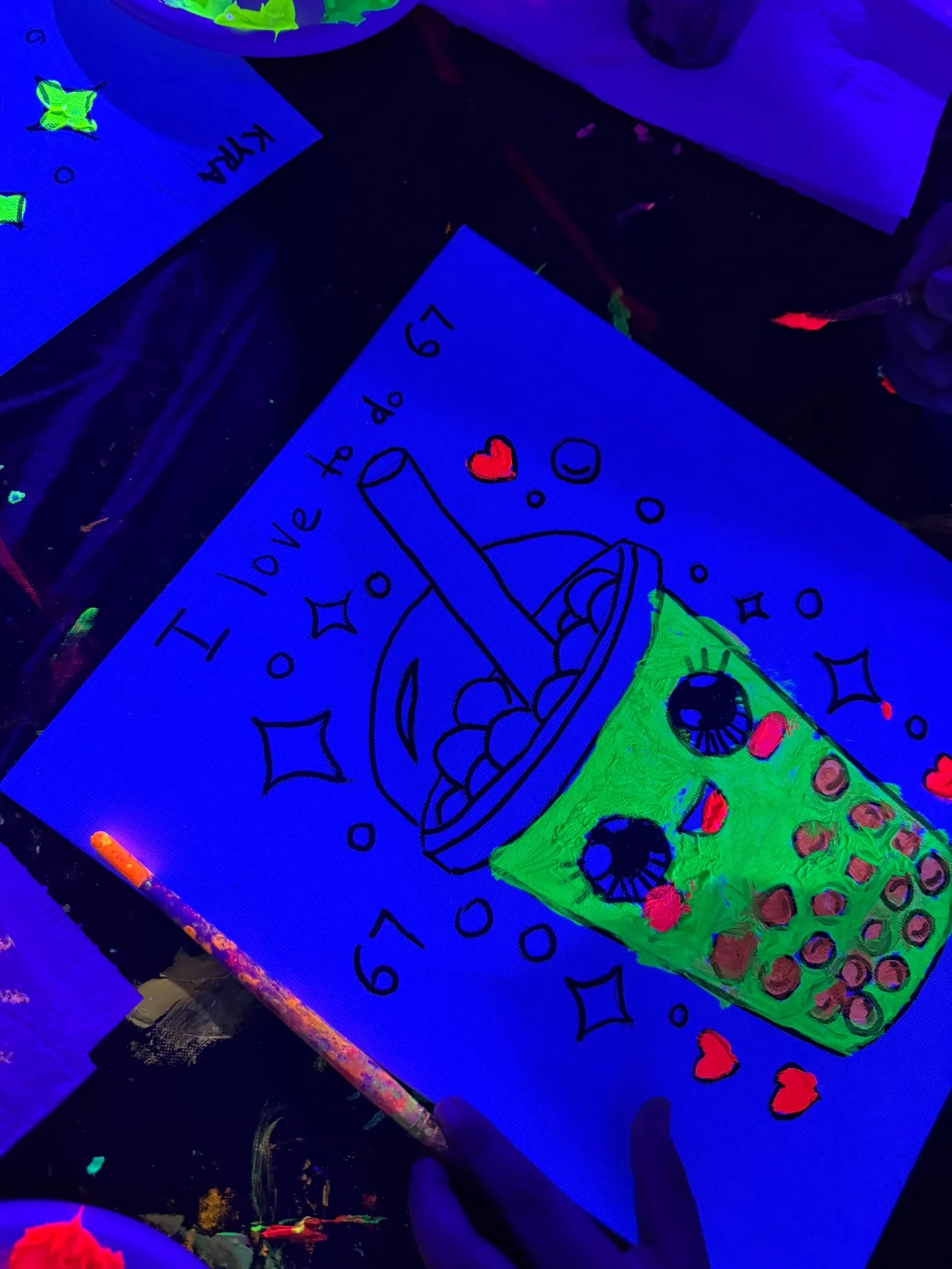 People holding colorful, neon, glow-in-the-dark artwork with various designs including animals, flowers, and abstract patterns, in a room decorated with glowing hearts, stars, and celestial shapes.
