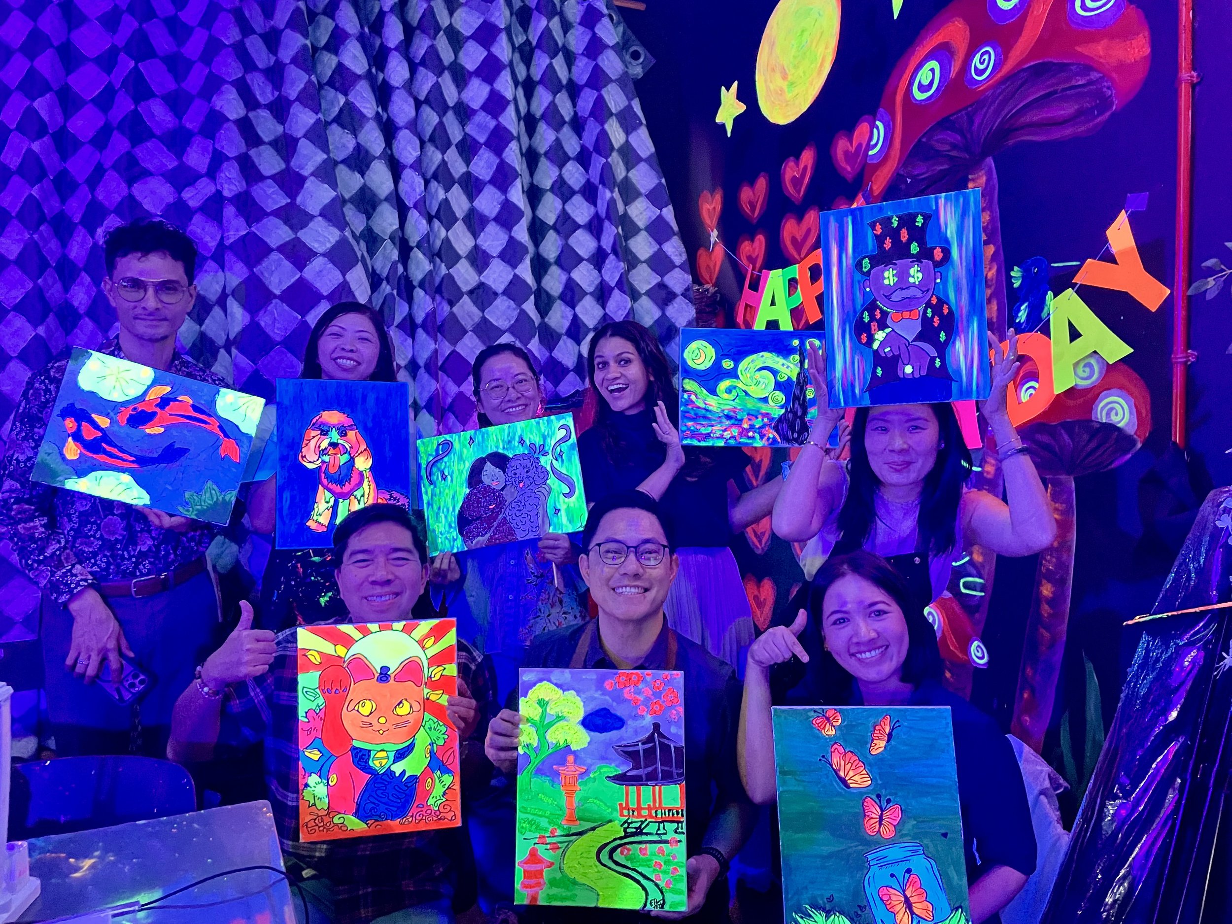 People holding colorful, neon, glow-in-the-dark artwork with various designs including animals, flowers, and abstract patterns, in a room decorated with glowing hearts, stars, and celestial shapes.