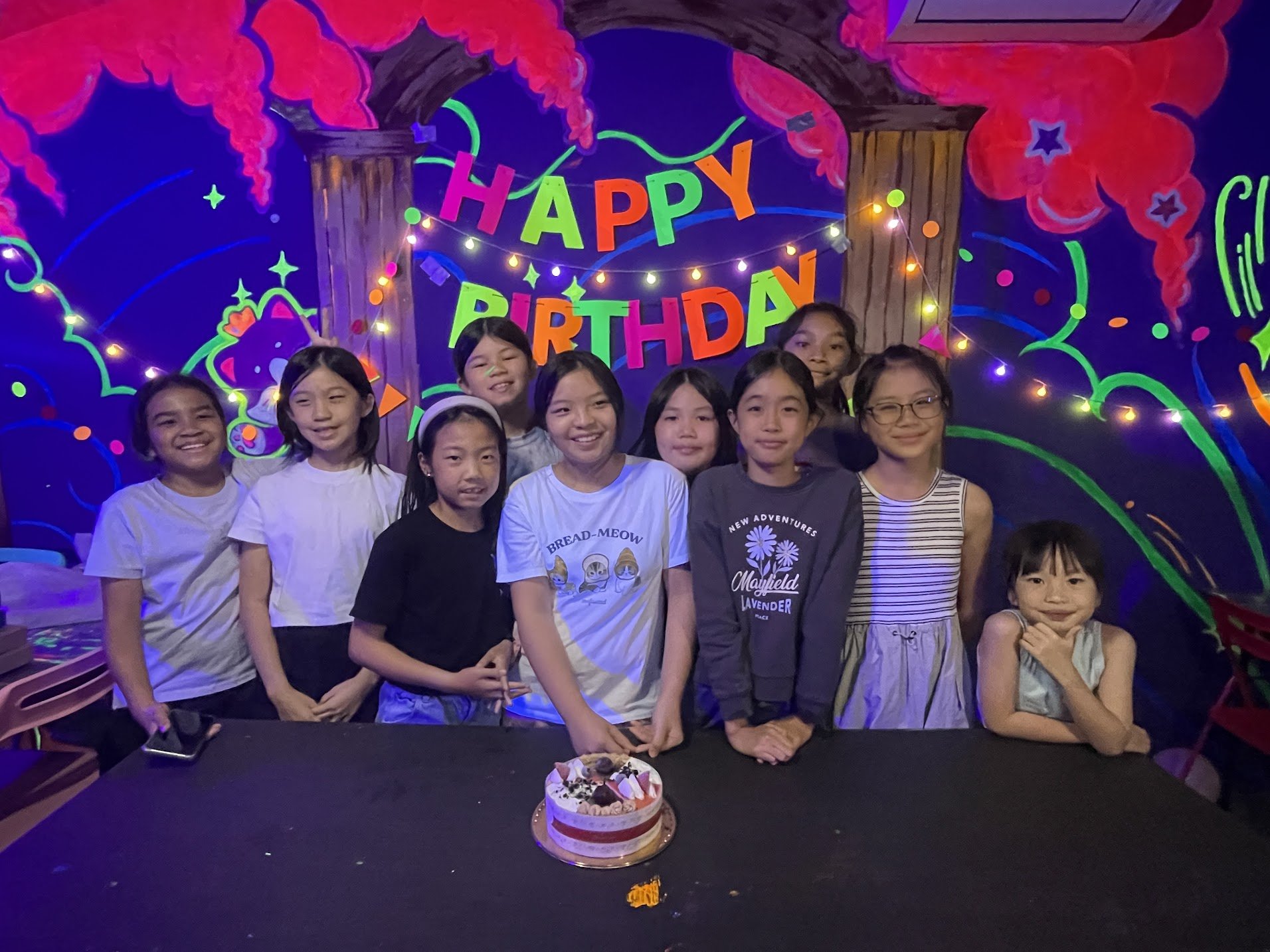 Group of nine smiling children standing behind a table with a birthday cake, with a colorful, decorated birthday party backdrop that says 'HAPPY BIRTHDAY' and vibrant decorative elements.