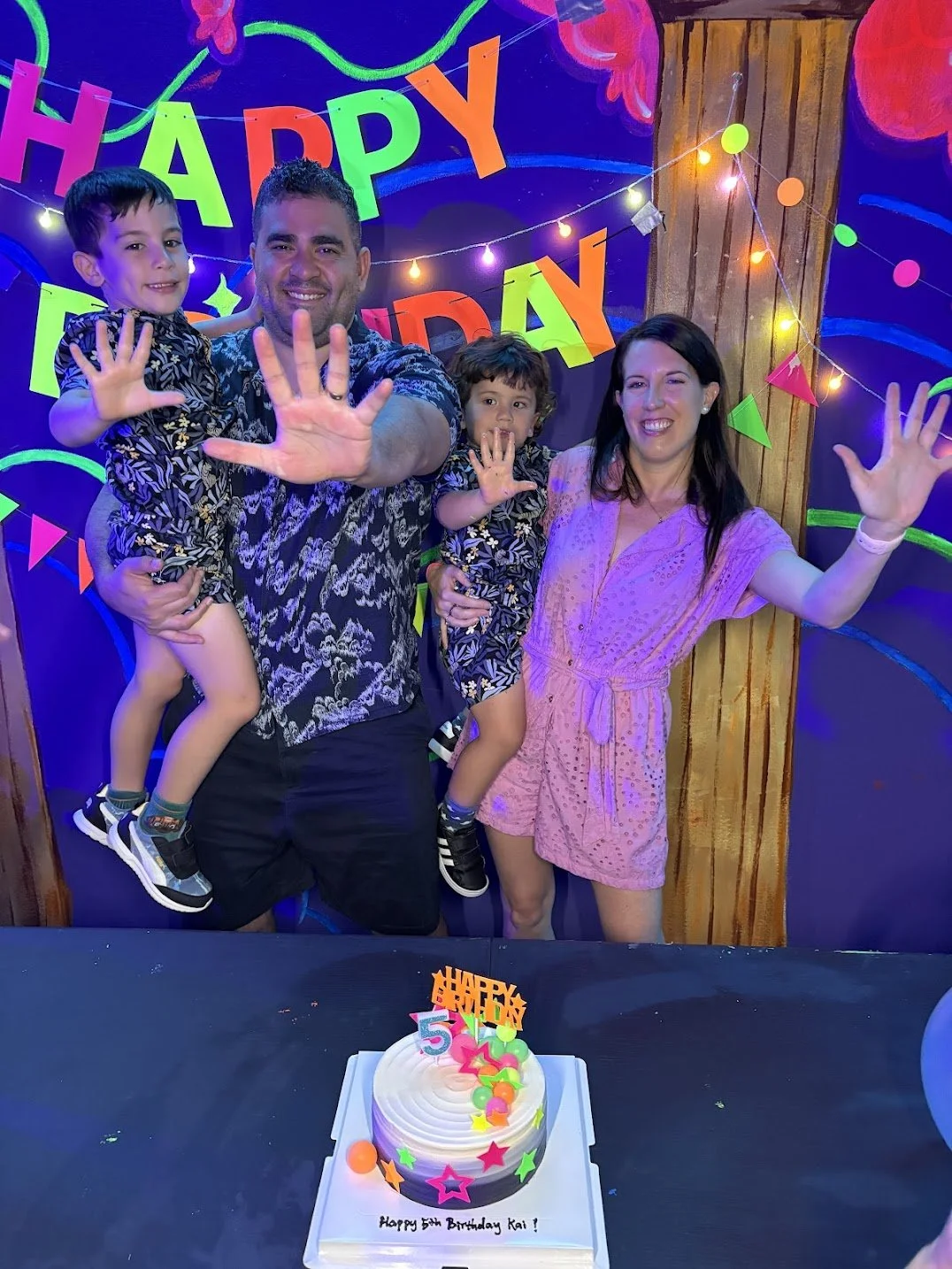 Family celebrating a child's 5th birthday with a cake, colorful decorations, and a 'Happy Birthday' banner in the background.