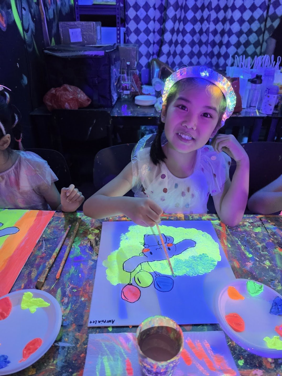 Girl with a glowing headband creating a neon-colored painting of a cartoon character on paper at a glow-in-the-dark art event.