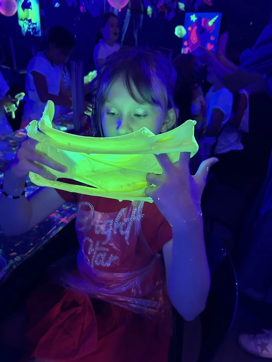 A young girl at a glow-in-the-dark party holding a fluorescent slime object, with other children and glowing artwork in the background.