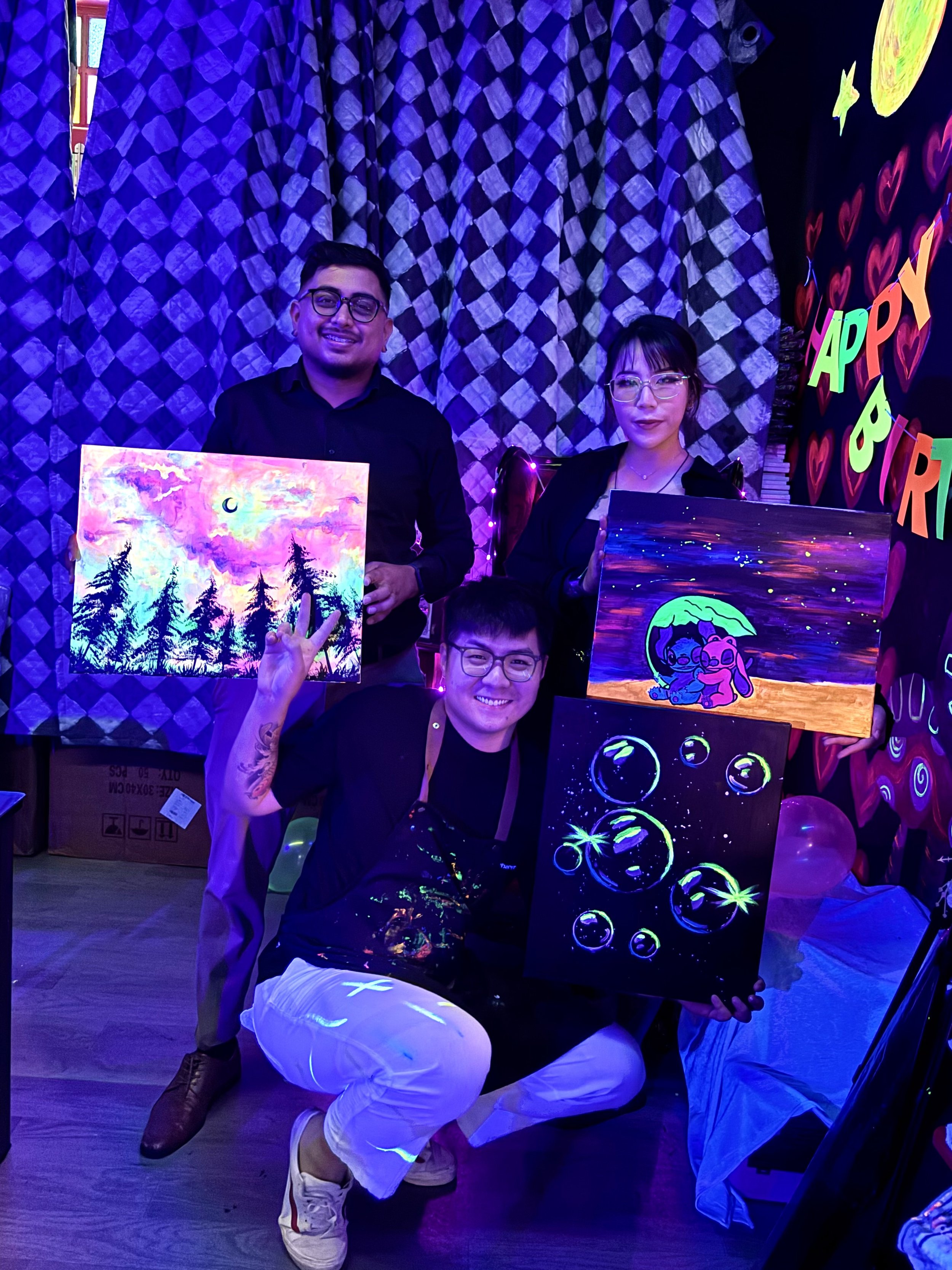 People holding colorful, neon, glow-in-the-dark artwork with various designs including animals, flowers, and abstract patterns, in a room decorated with glowing hearts, stars, and celestial shapes.