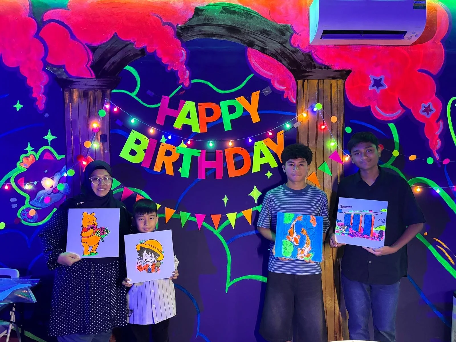 Four people standing in front of a colorful 'Happy Birthday' banner with neon decorations. They are holding drawings and standing in a decorated room with neon art and string lights.