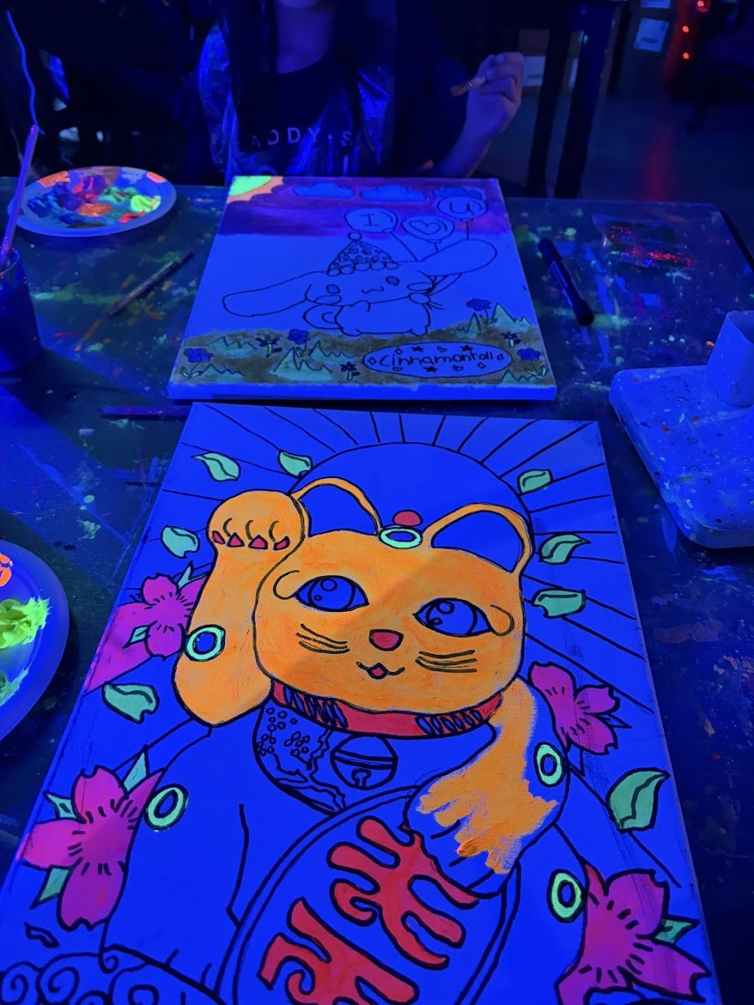 Glow-in-the-dark paintings of a cartoon orange cat with a hand on its face, surrounded by pink flowers and green leaves, illuminated by blacklight.