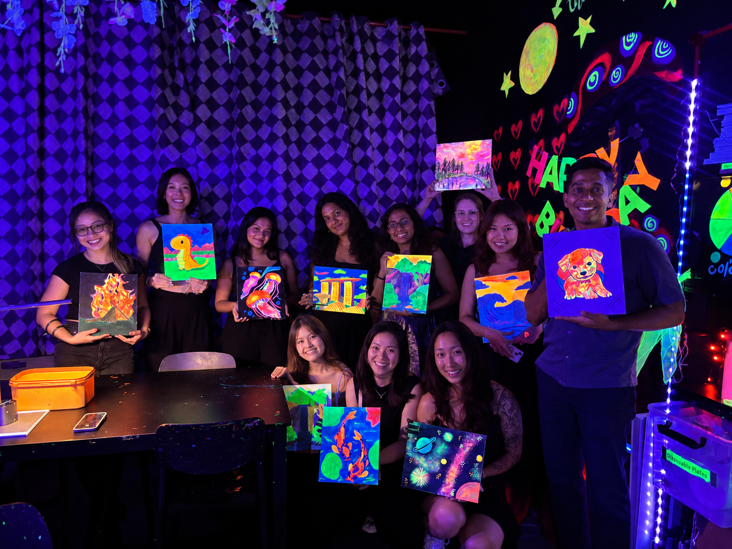 People holding colorful, neon, glow-in-the-dark artwork with various designs including animals, flowers, and abstract patterns, in a room decorated with glowing hearts, stars, and celestial shapes.