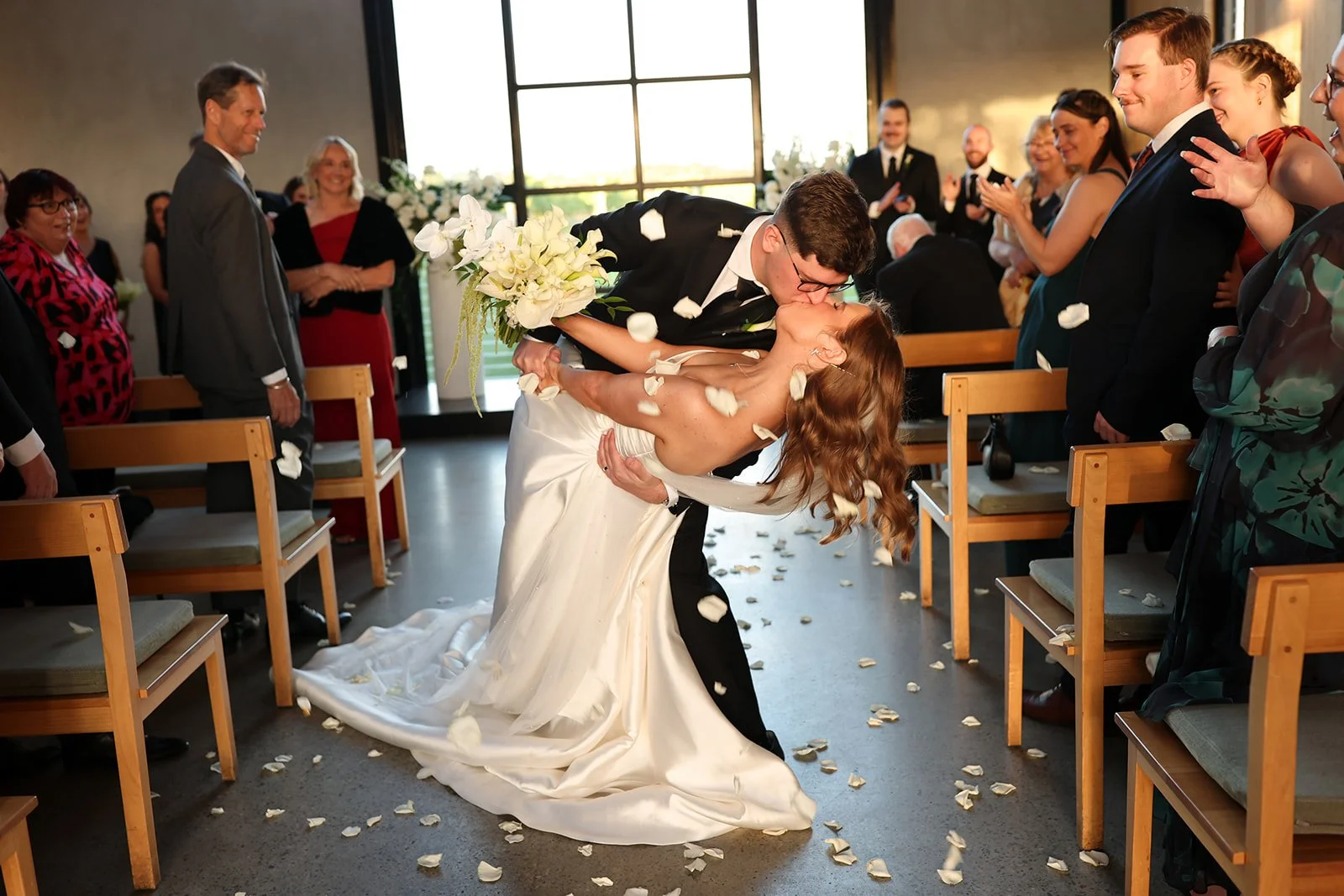A bride and groom kissing during their wedding ceremony in a church, with guests standing and smiling, petal-covered floor, and large windows letting in natural light.