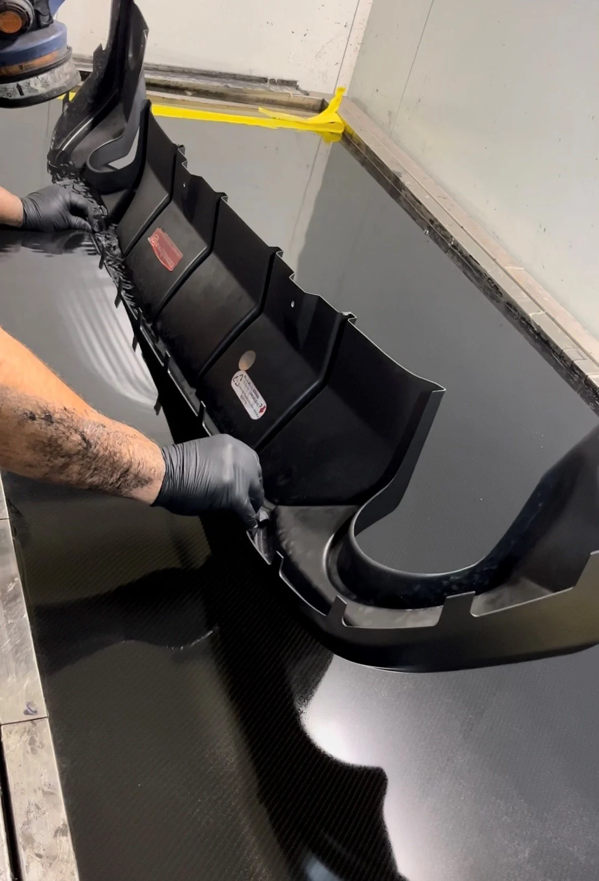 Person hydro dipping a car bumper diffuser with gloved hands in a workshop. Hydrographics WA, Western Australia's premier hydro dipping specialist