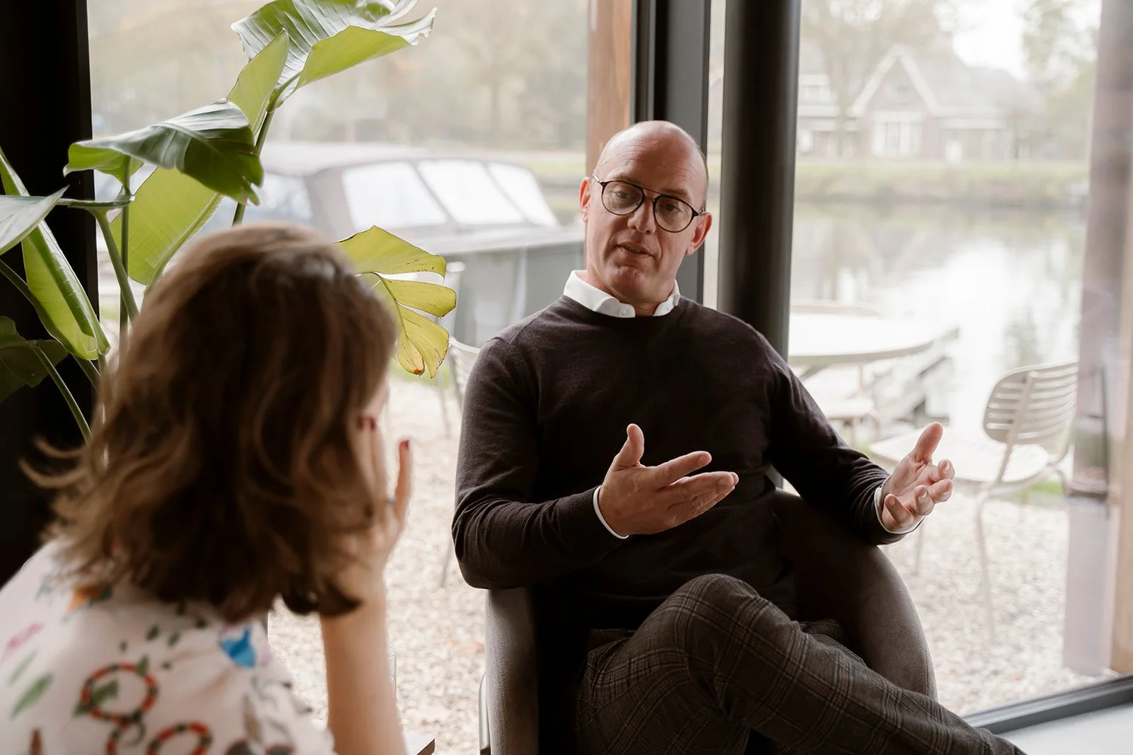 Een man met overhemd en bruine trui zit in een vergaderlocatie en probeert een vrouw te overtuigen. Het is een zakelijk gesprek.