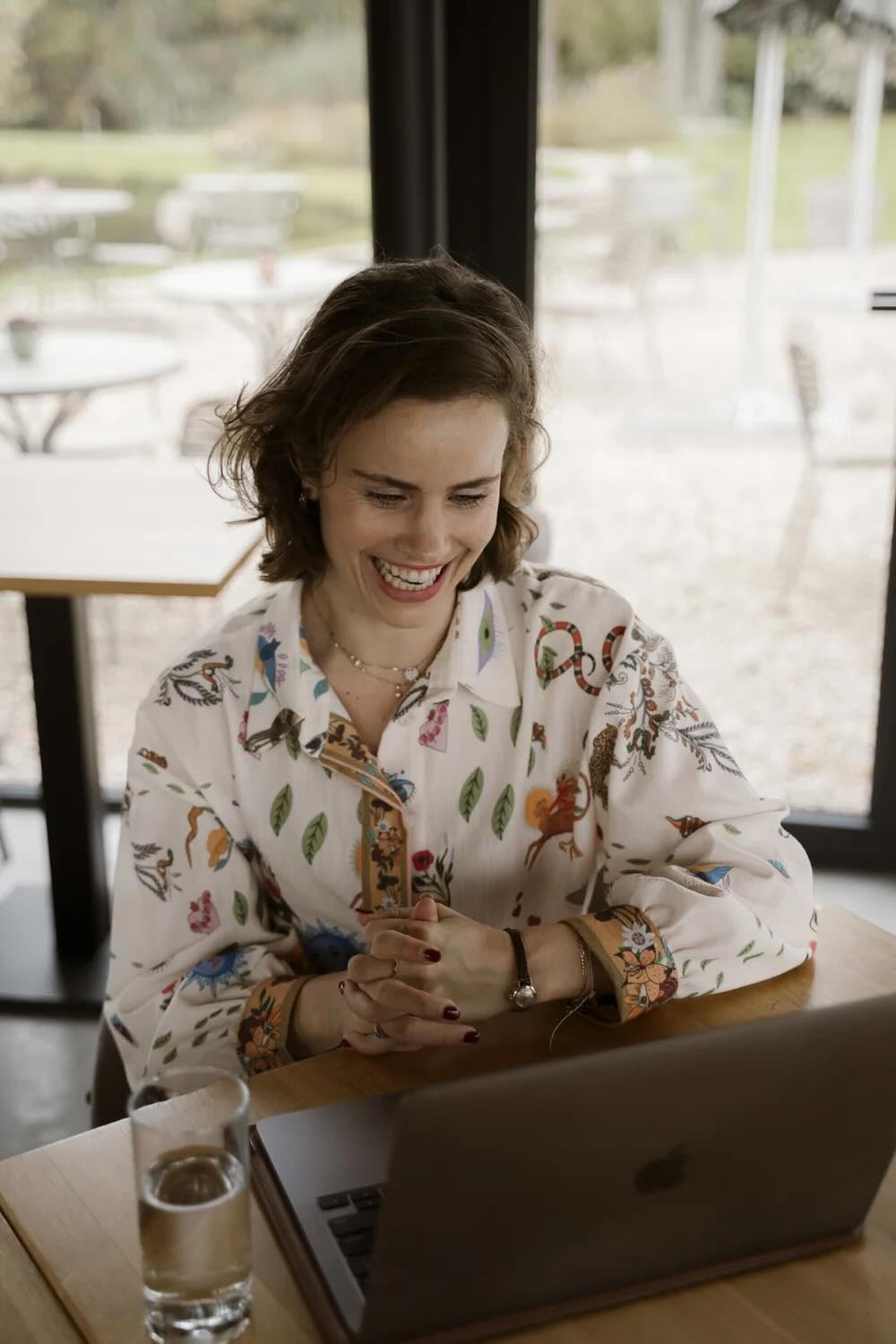 Een vrouw, Lindy Bremer, zit aan een houten tafel in een ruimte met grote ramen, kijkt naar een laptop en glimlacht tijdens een digitaal kennismakingsgesprek met een client.