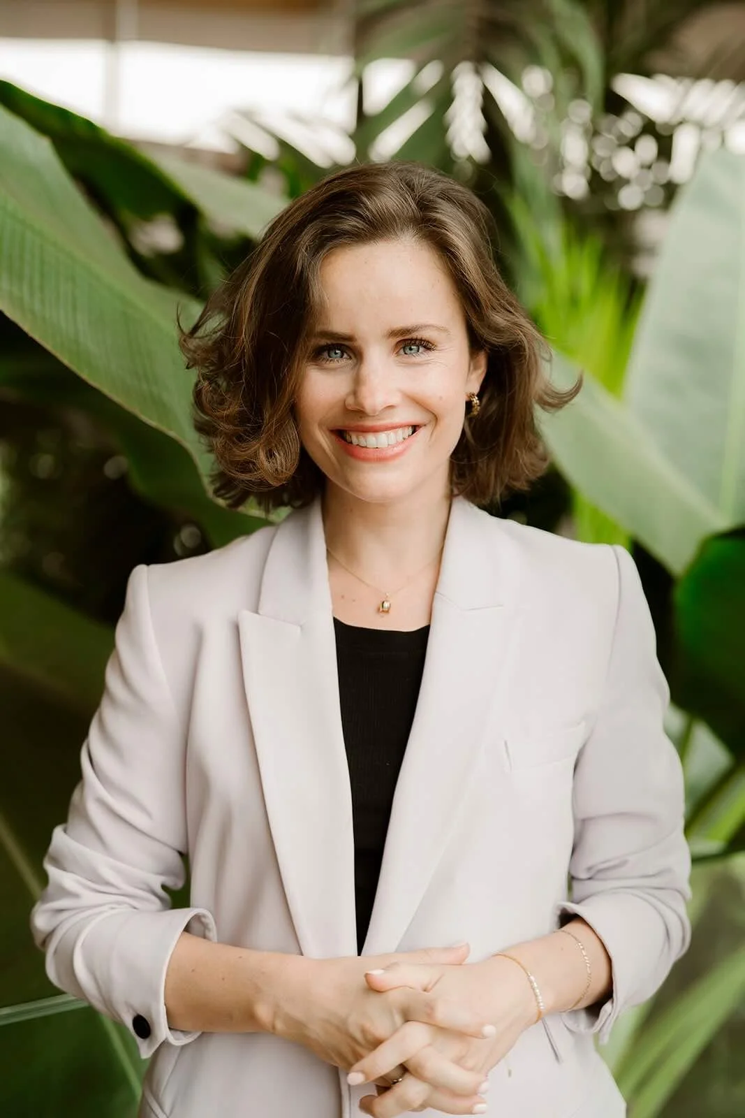 Lindy Bremer in lichtgekleurde blazer glimlacht terwijl ze voor grote groene bladeren staat, handen in elkaar gevouwen.