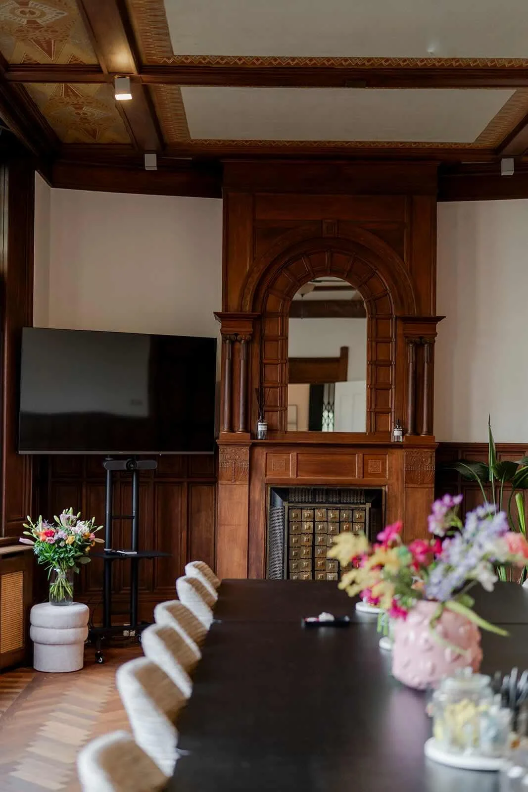 Een elegante vergaderruimte met een lange tafel in Den Haag, omringd door twintig gestoffeerde stoelen, met een grote vaas met bloemen en een antieke houten open haard.