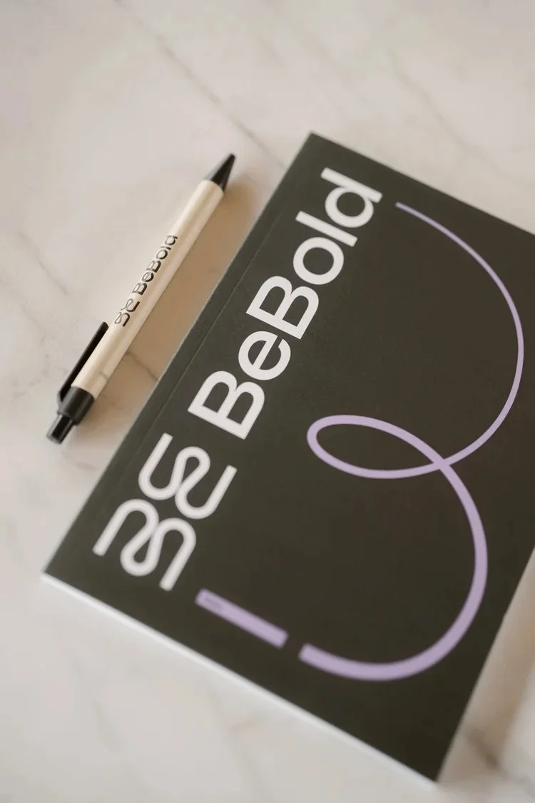 Een notitieboek van BeBold Academy met witte en paarse tekst en een pen liggen op een witte marmeren tafel.